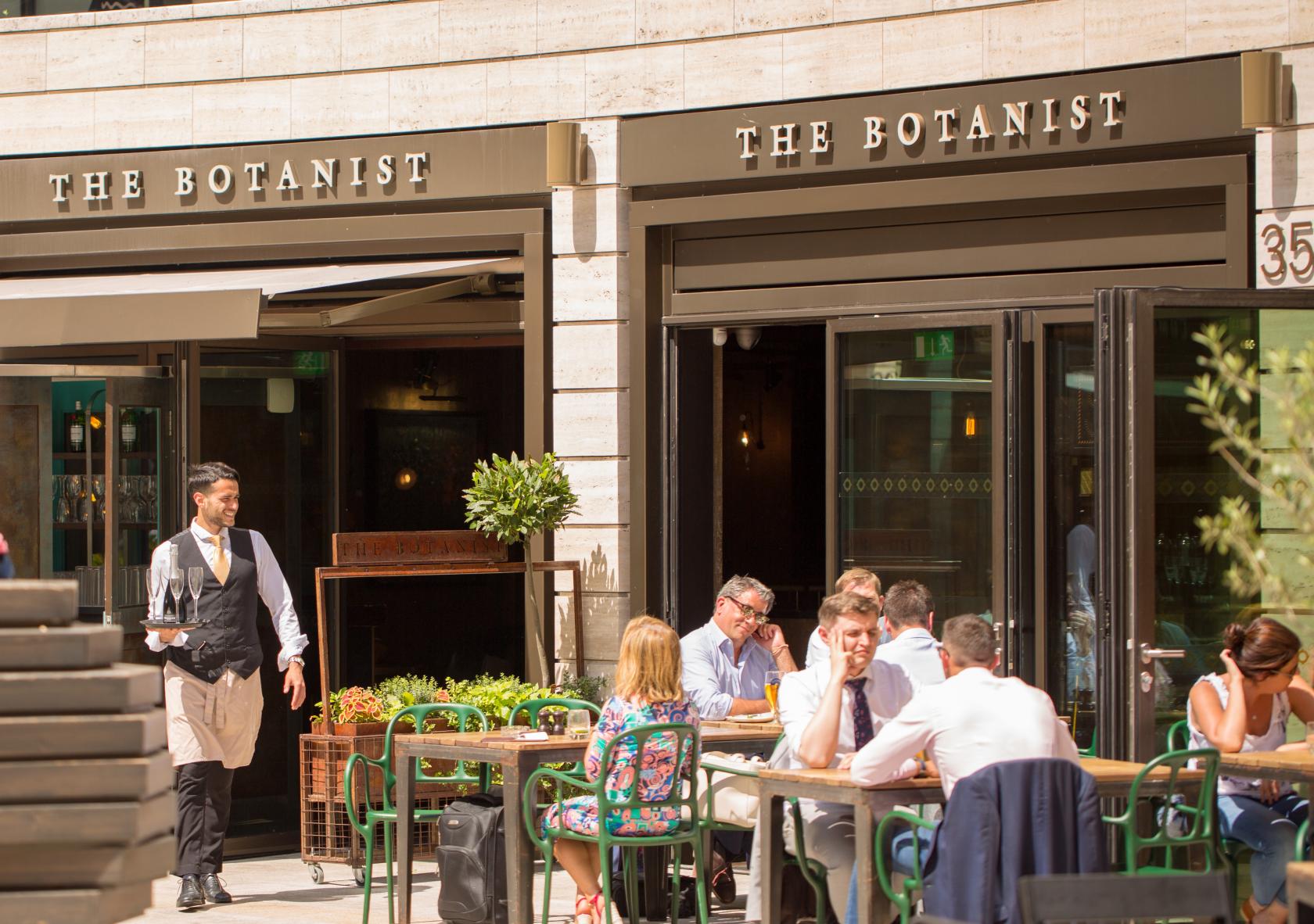 An outdoor restaurant seating area with wooden tables, green chairs, and patrons dining at The Botanist Broadgate Circle.