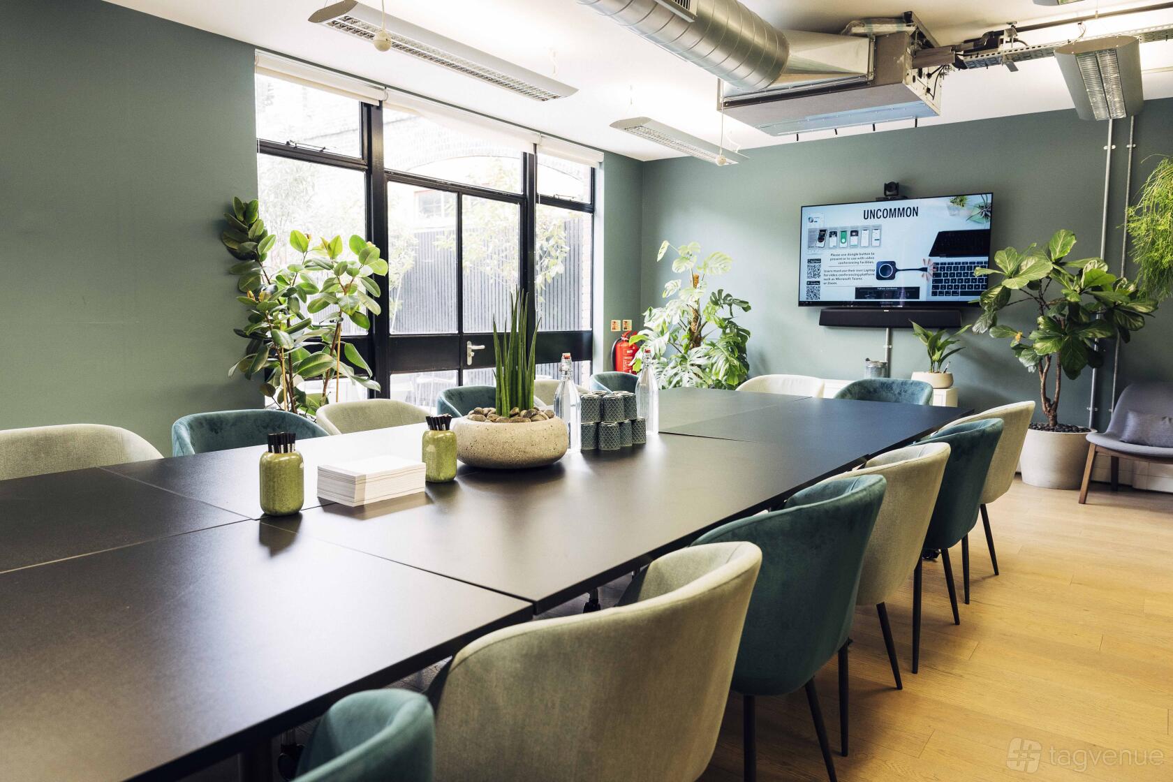 A meeting centre with large windows, green velvet chairs, potted plants, and a wall-mounted screen at Uncommon Fulham.