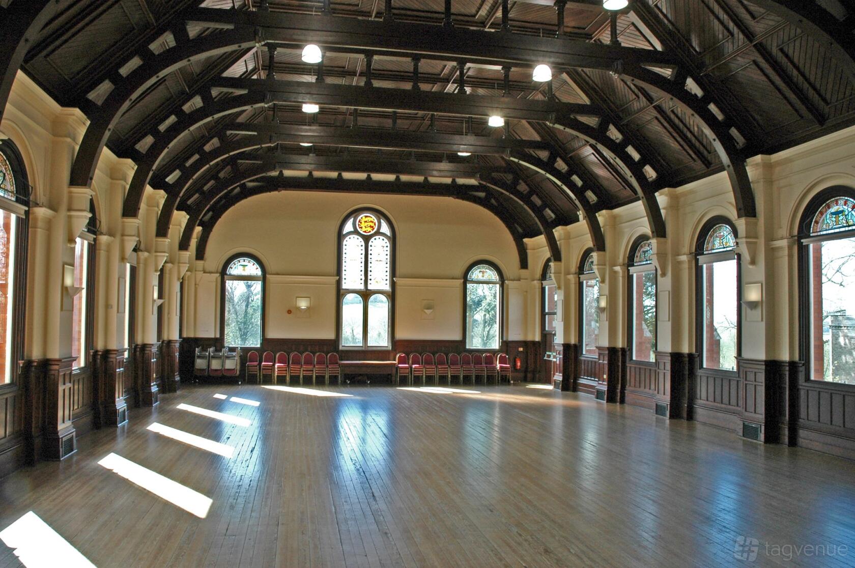 A spacious ballroom with arched wooden beams, tall arched windows, and polished wood floors at Charlton Down Village Hall.