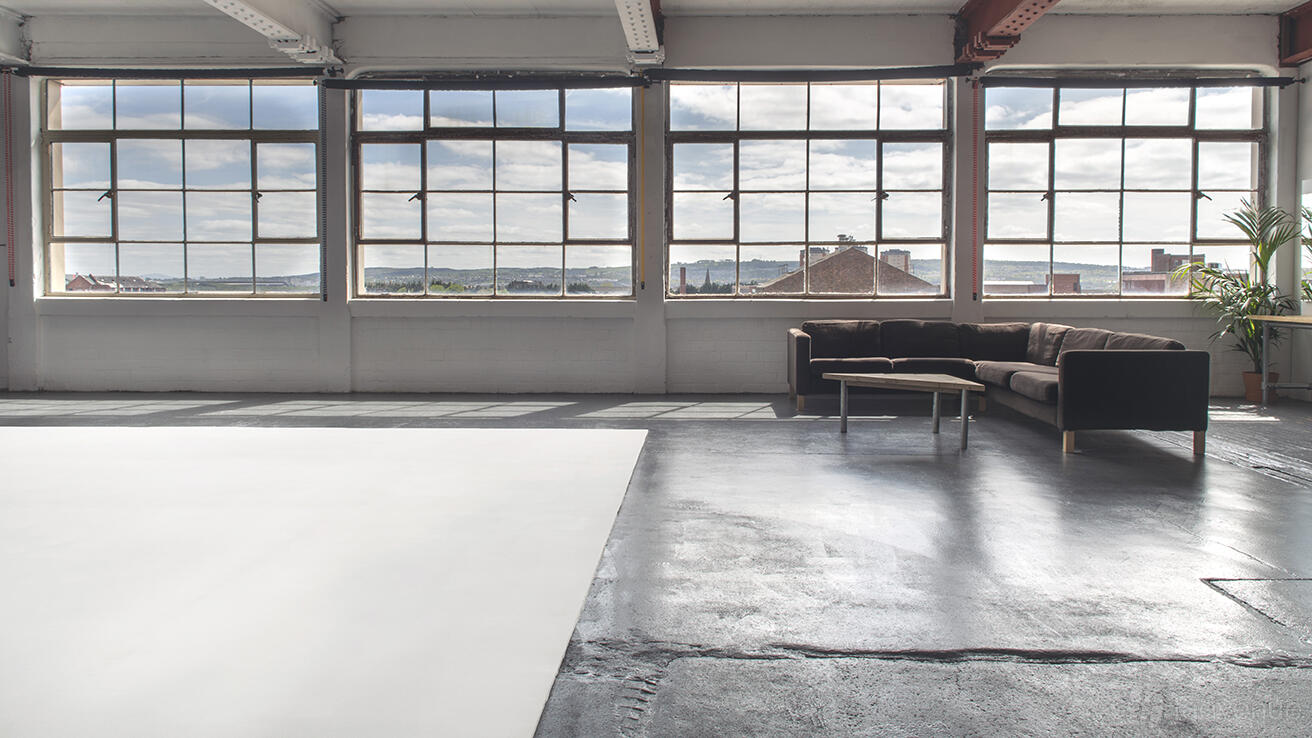 A photo studio with large industrial windows, concrete floors, and a lounge area with a brown sofa at Broadscope Studios.