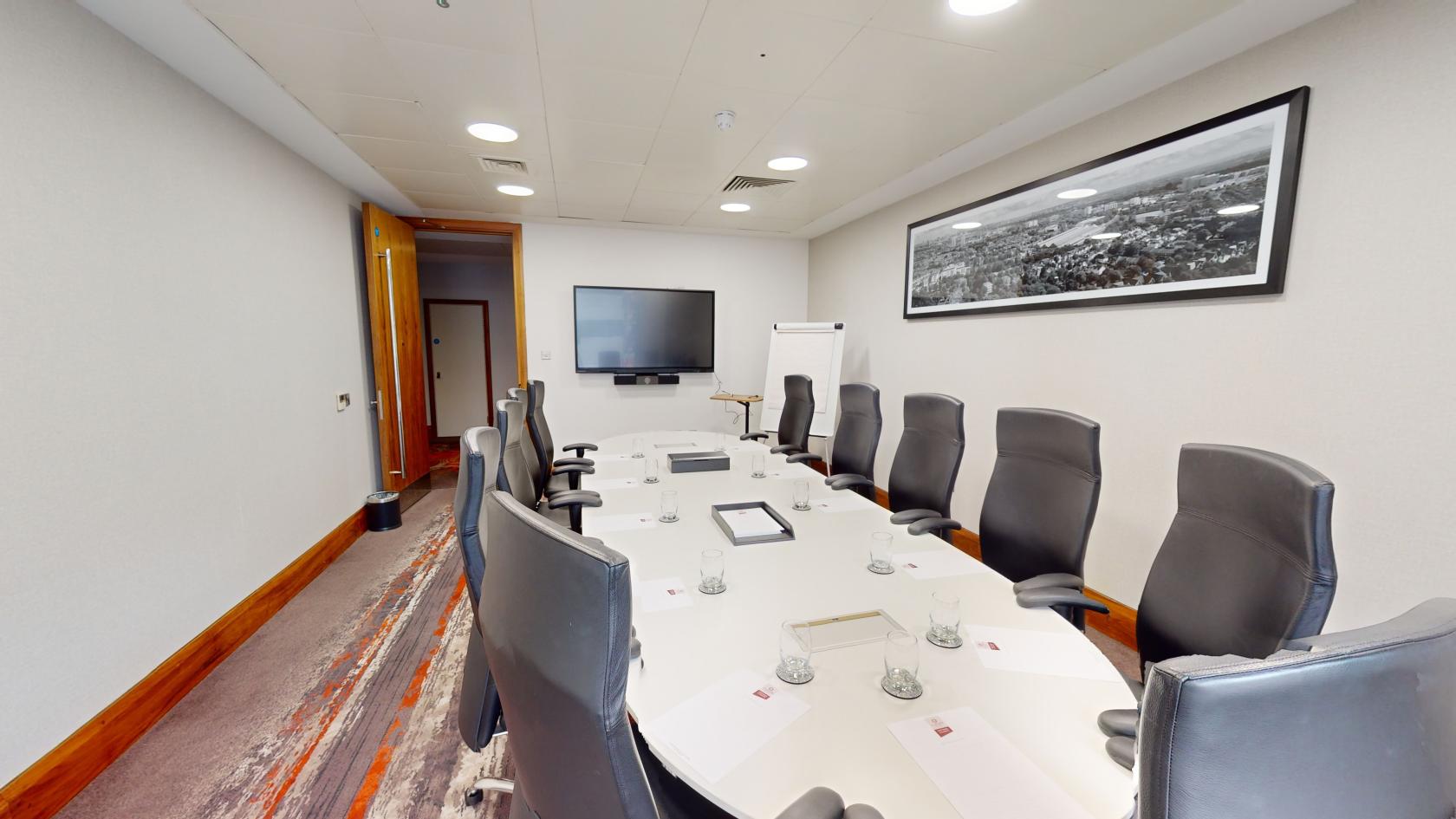 A hotel meeting room with a long oval table, black executive chairs, wall-mounted TV, and a panoramic city photo at Clayton Hotel Birmingham.