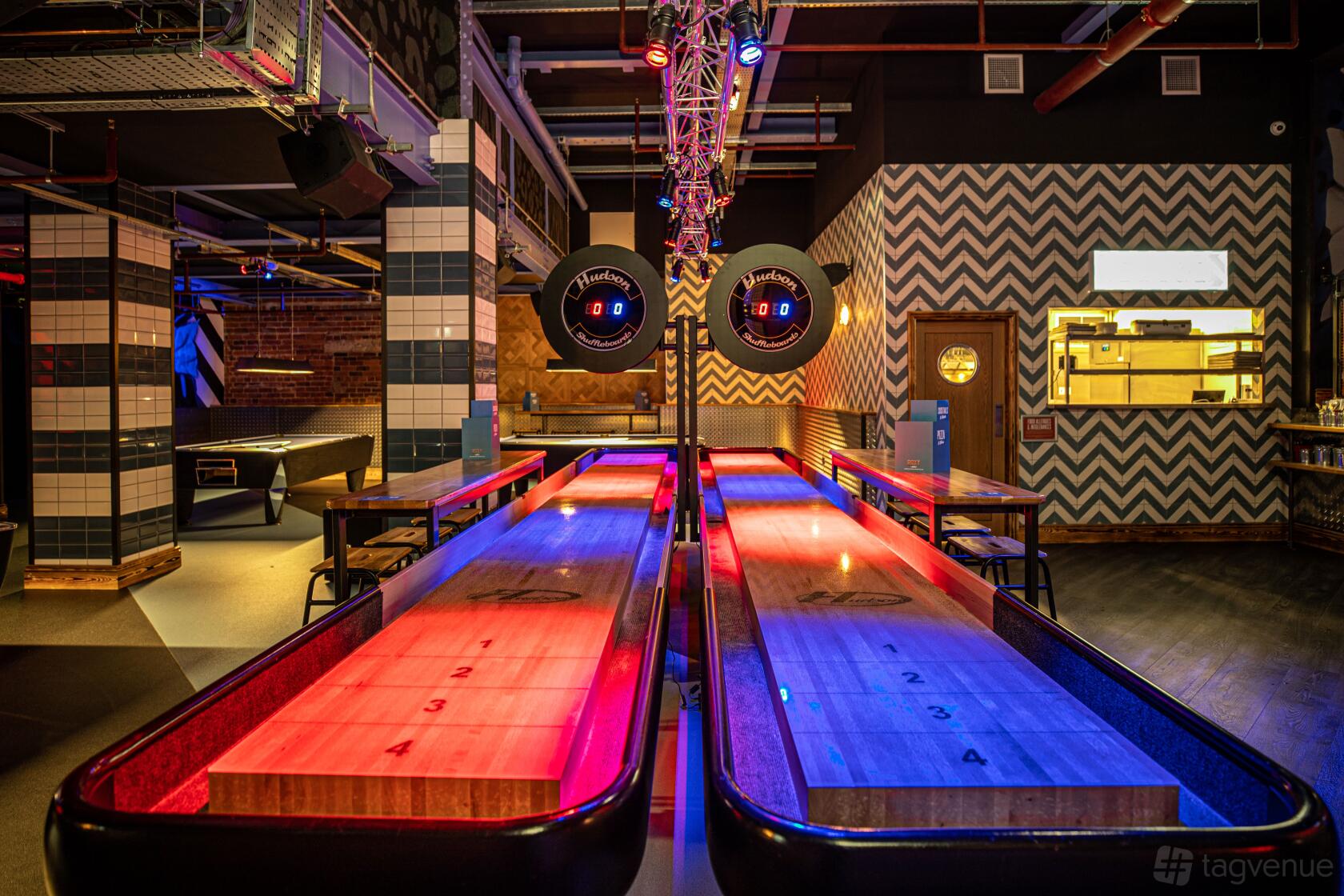 An event space in a bar with illuminated shuffleboard tables, zigzag patterned walls, and overhead scoreboards at Roxy Lanes Leeds The Light.