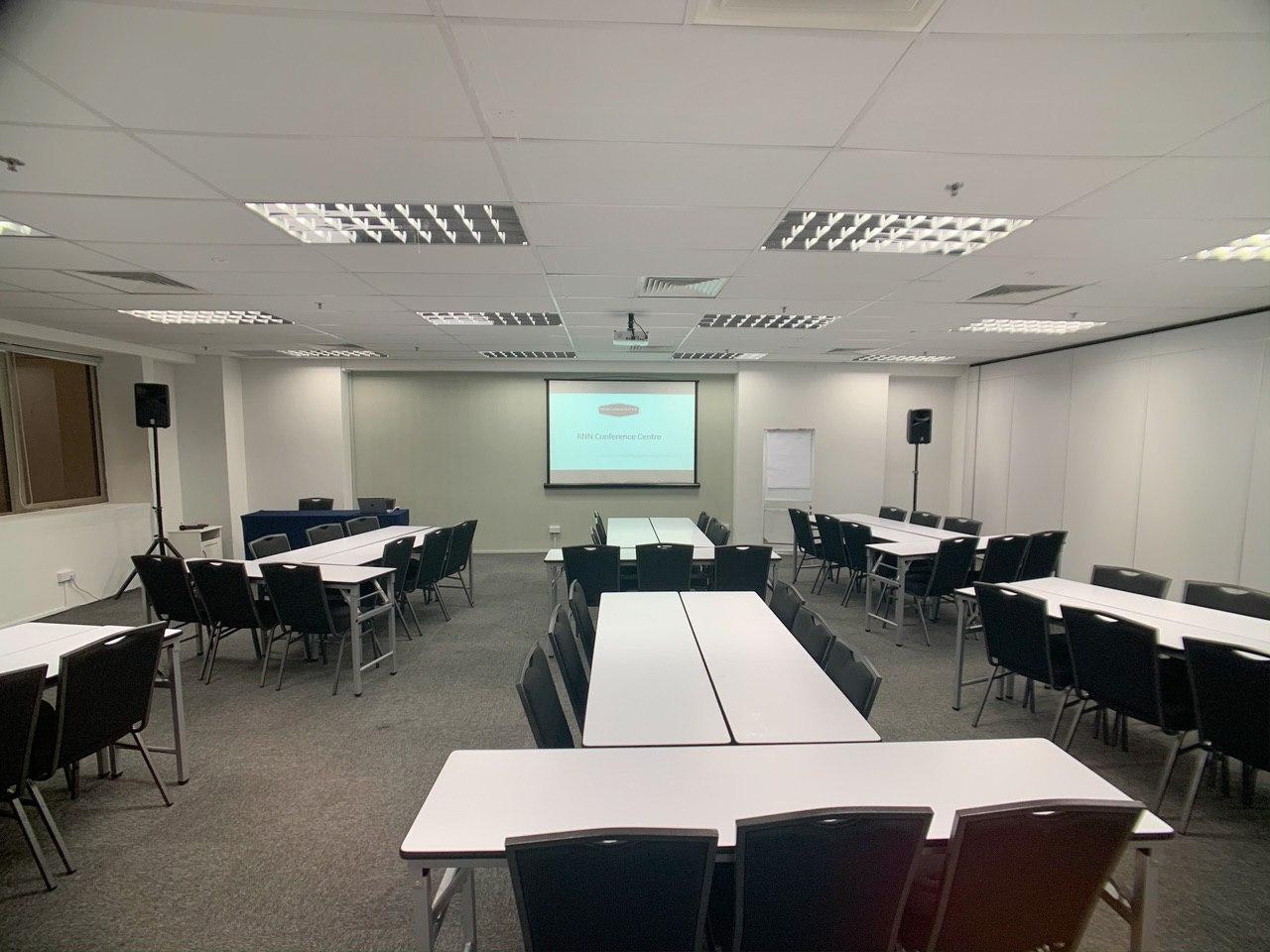 An auditorium with arranged conference tables, black chairs, and a projector screen at RNN Conference Centre.