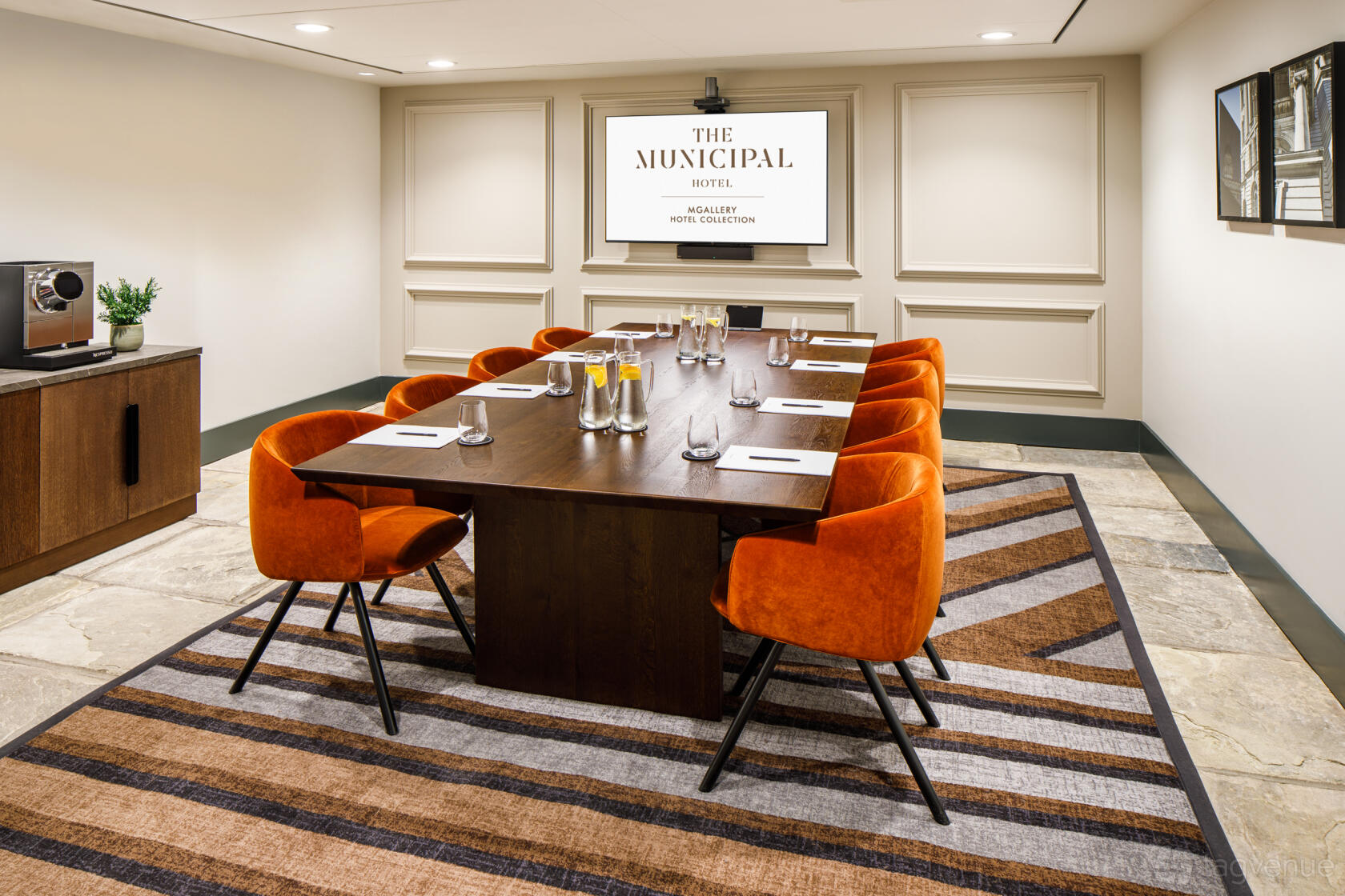 A meeting room with a long wooden table, orange chairs, wall screen, and striped rug at Municipal Hotel Liverpool - MGallery.