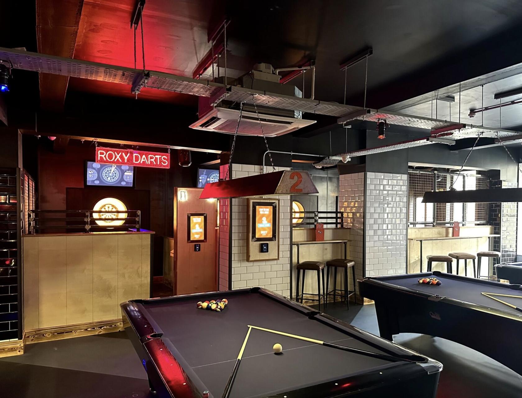 An event space in a bar with pool tables, bar stools, and neon darts signage at Roxy Ball Room Leeds Boar Lane.