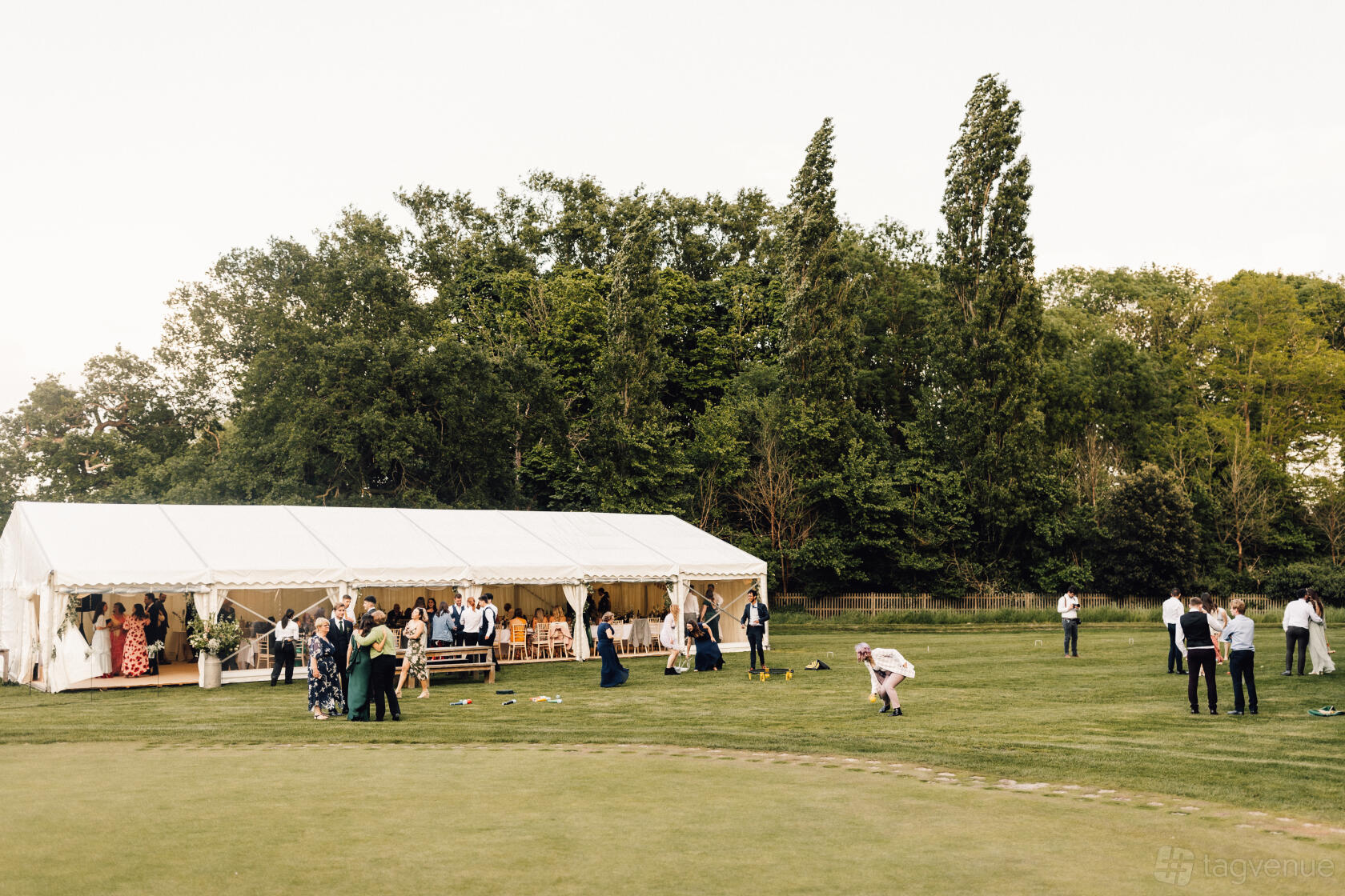An outdoor event space with a white marquee tent on green lawns surrounded by trees at Hampton Court Palace Golf Club.