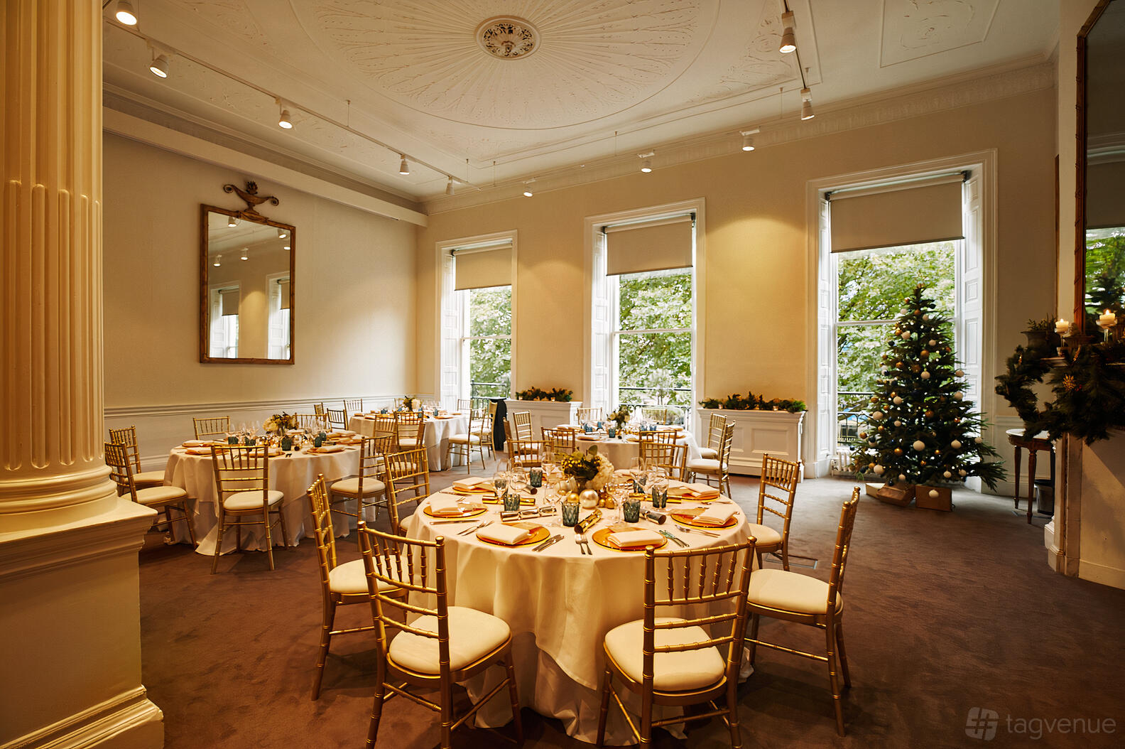 An event venue with round tables covered in white linens, gold chairs, large windows, and a decorated Christmas tree at 41 Portland Place.