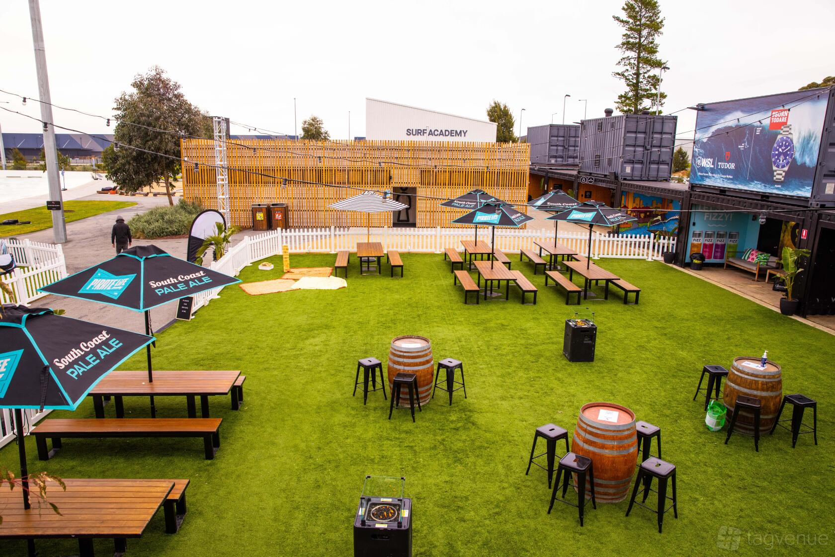 An outdoor event venue with picnic tables, barrel tables, and branded umbrellas on artificial grass at URBNSURF.