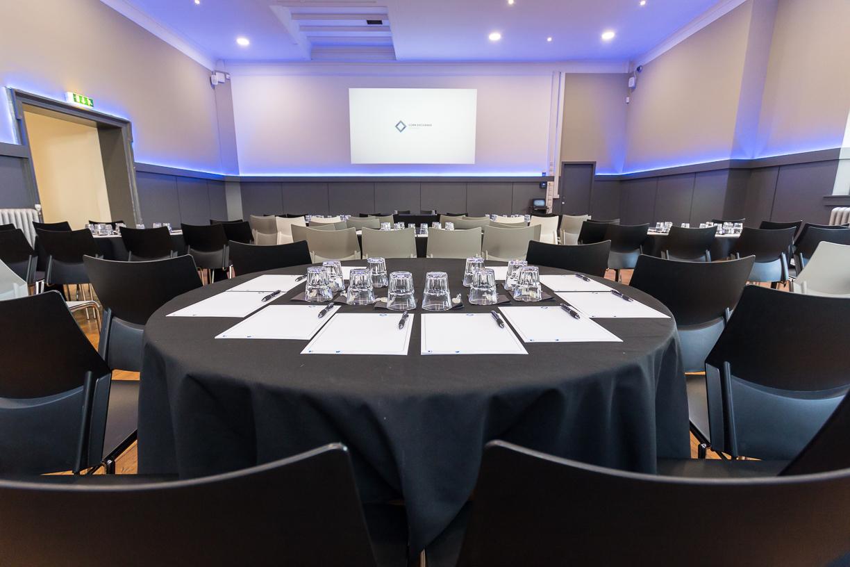 An event room with round tables set with notepads, glasses, and rows of chairs facing a projector screen at Edinburgh Corn Exchange.