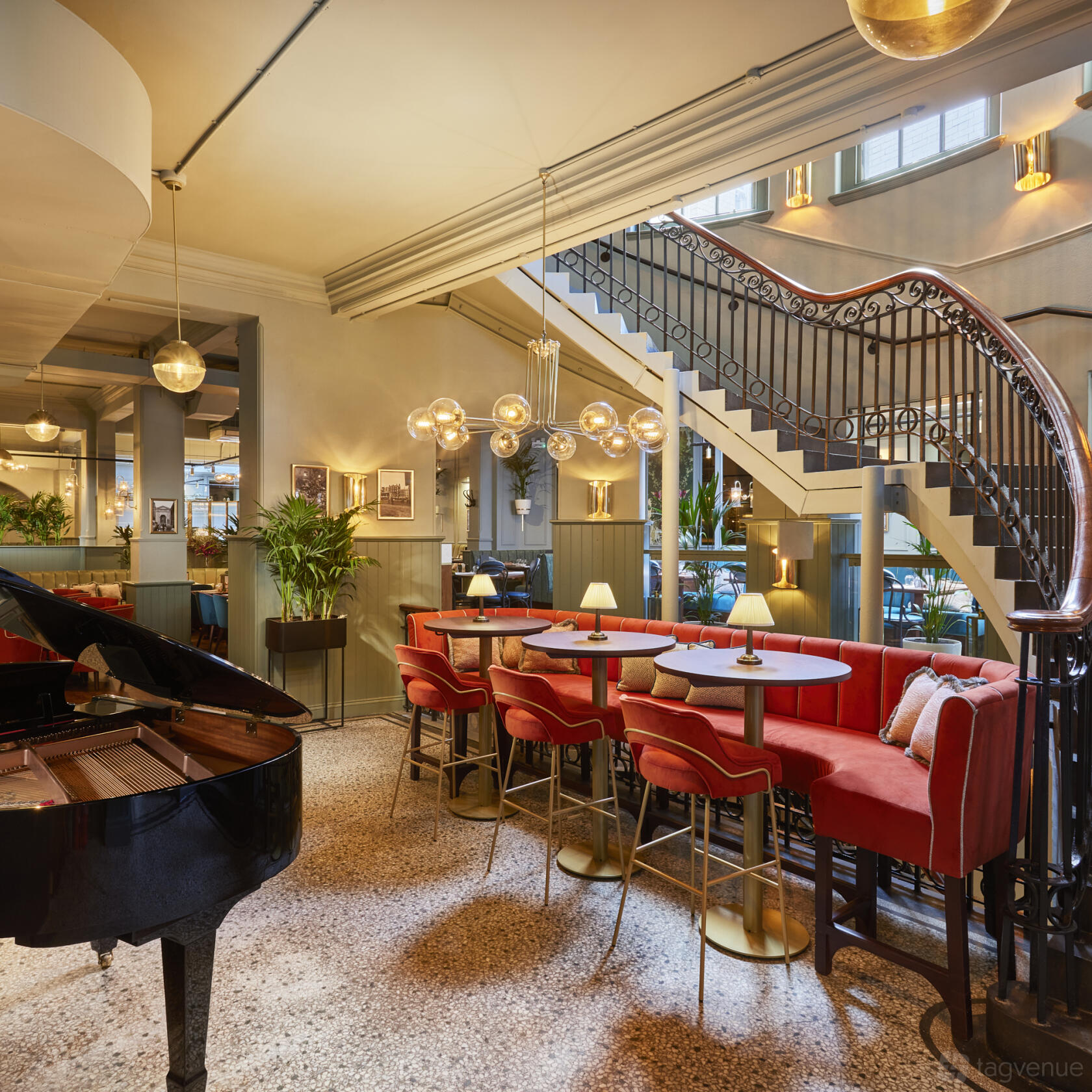 A restaurant with round tables, red upholstered seating, a grand piano, and a curved staircase at Browns Covent Garden.