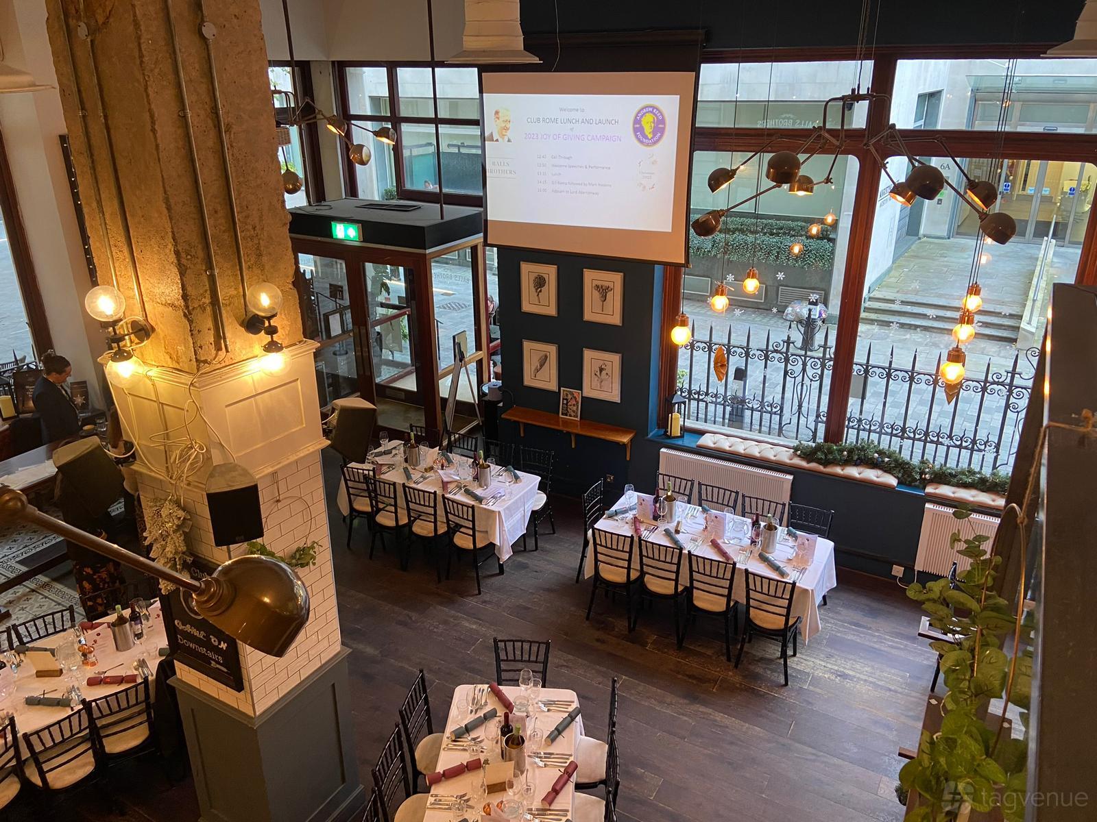 A bar with floor-to-ceiling windows, dark wood floors, and tables set with white linens at Balls Brothers Austin Friars.