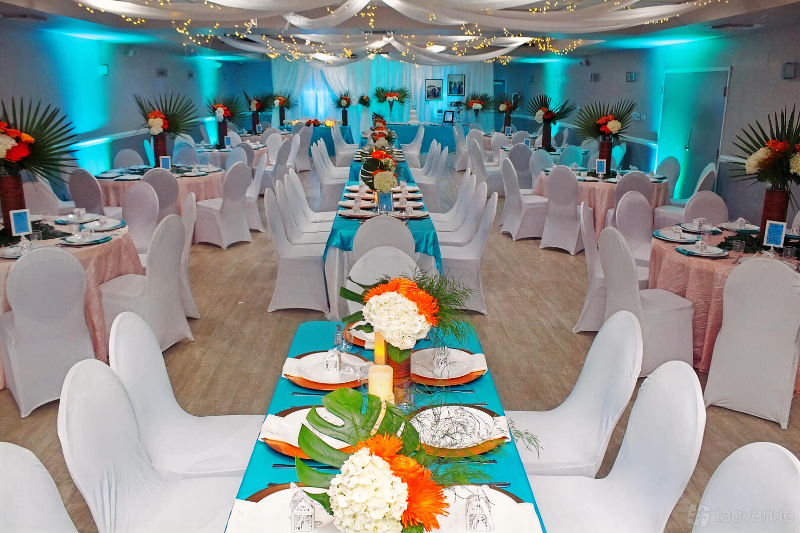 A banquet hall with turquoise table runners, tropical floral centerpieces, and draped ceiling lights at Tropical Paradise Banquet Hall.