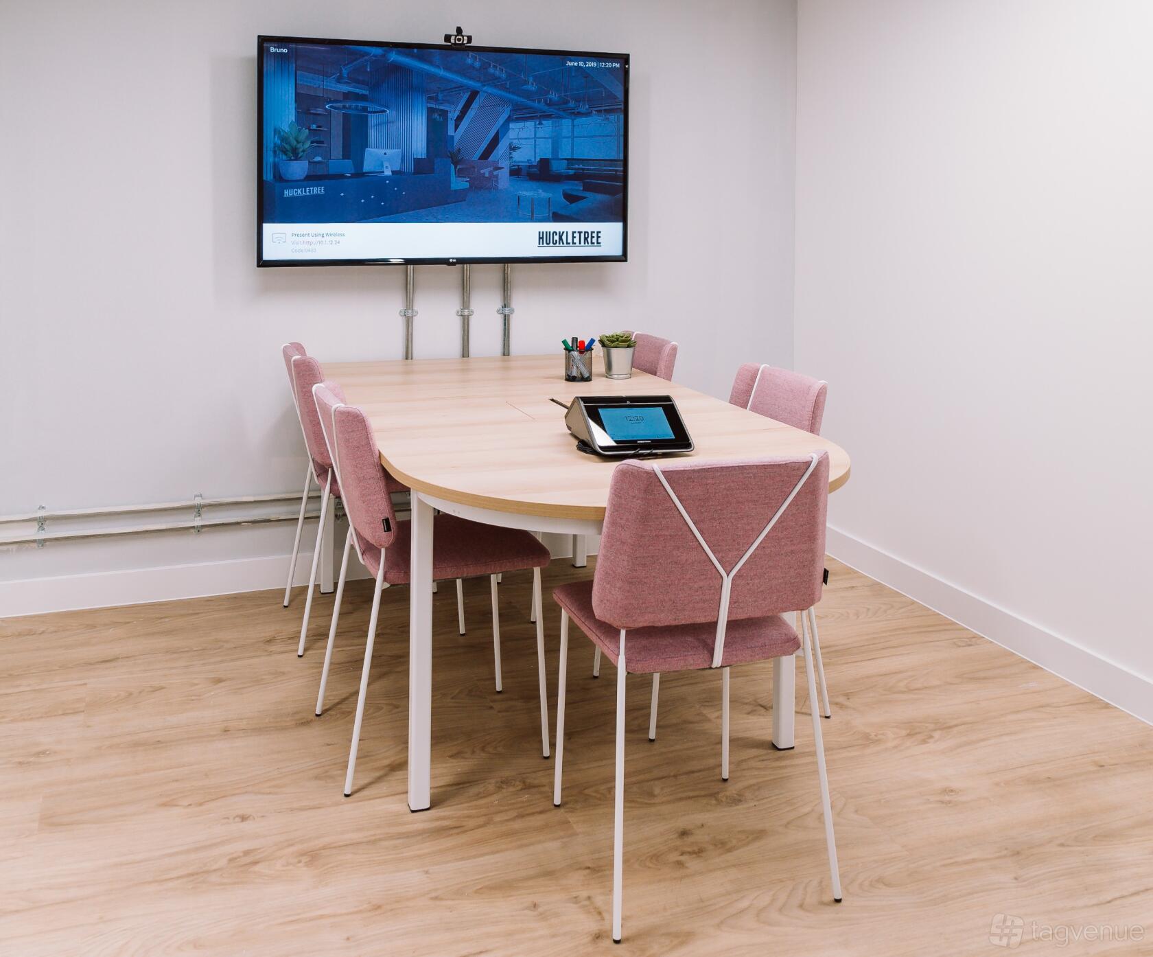 A meeting room with a wooden table, pink chairs, large wall-mounted screen, and tablet at Huckletree Soho.