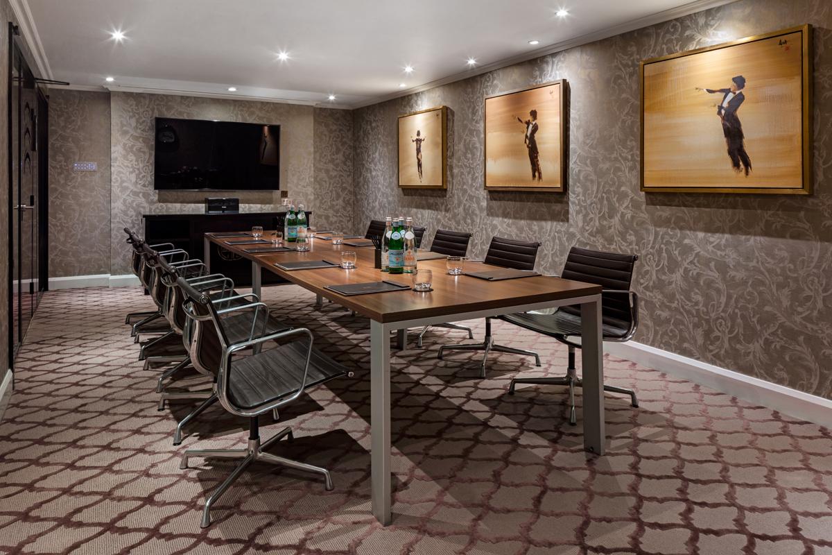 A hotel meeting room with a long wooden table, leather chairs, wall art, and a large TV screen at The May Fair Hotel.