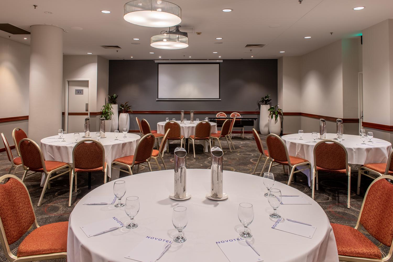 A function room with round tables, orange chairs, water glasses, and a projection screen at Novotel Perth Langley.
