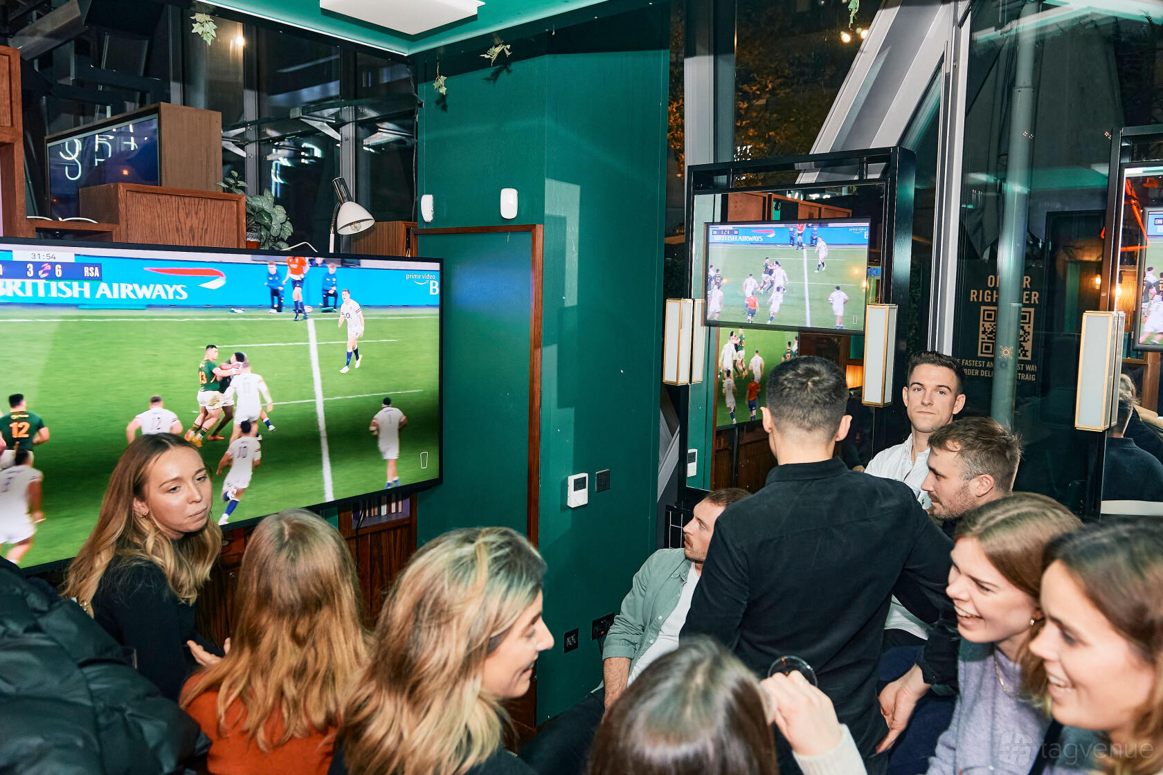 A pub bar with large screen televisions showing sports, green walls, and groups of people at Greenwood.