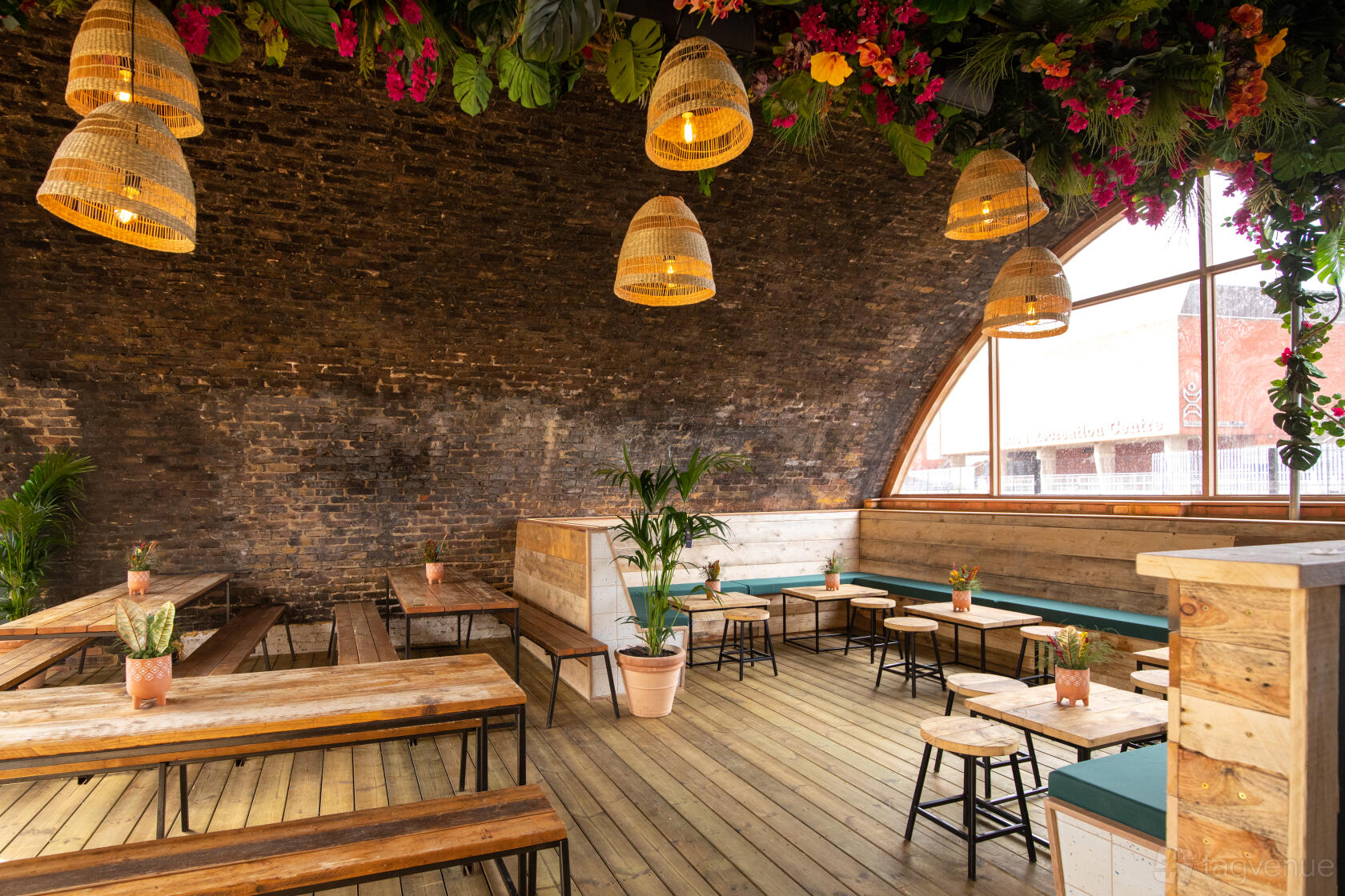 A bar with exposed brick arches, wooden benches, hanging basket lights, and leafy ceiling decor at Pergola Brixton.