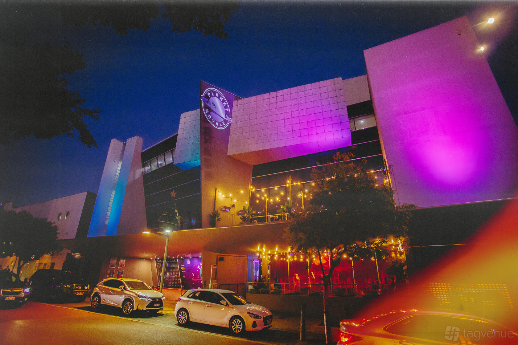 A pub with colorful exterior lighting, large windows, and an outdoor seating area at Planet Royale.