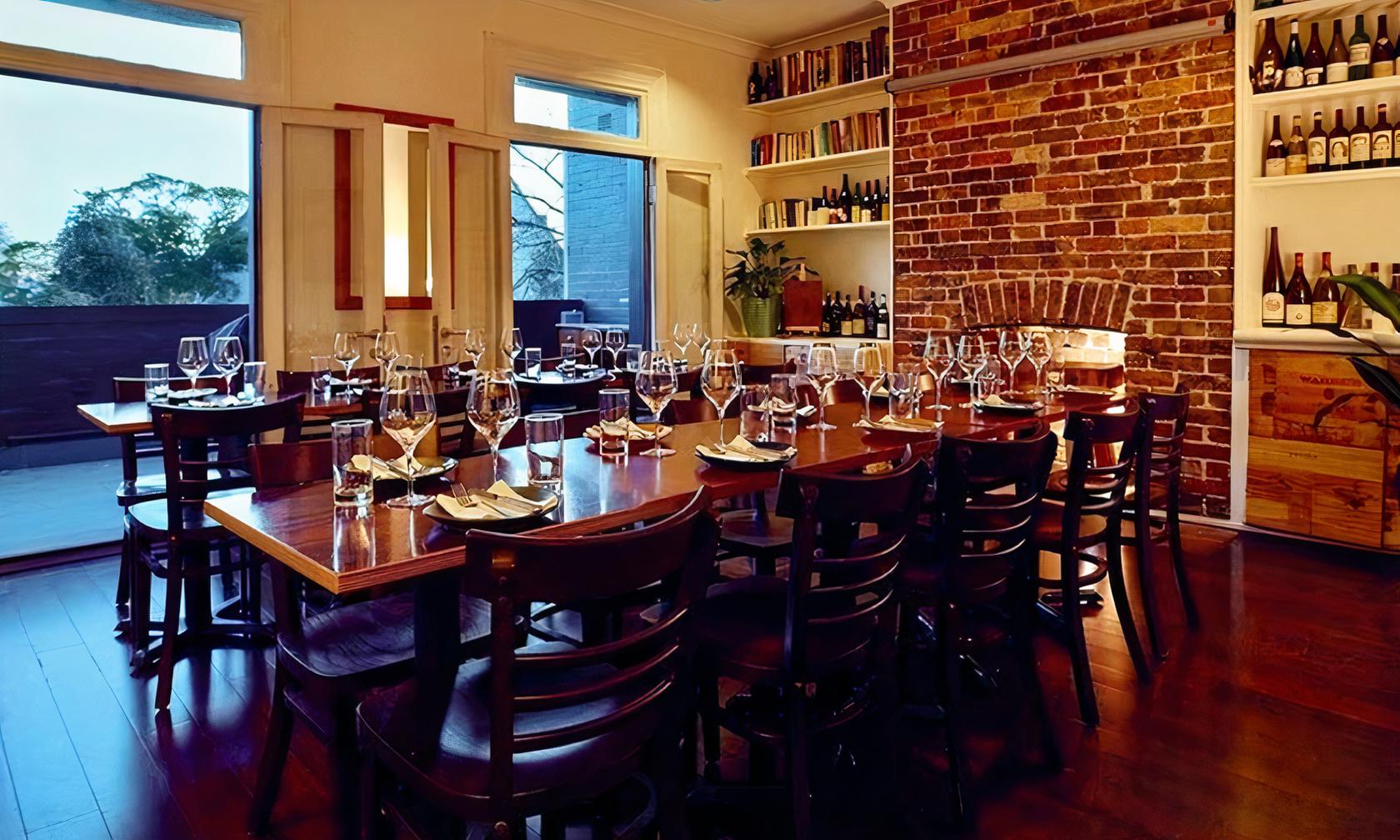 A dining room with exposed brick walls, wooden tables set with glassware, and built-in wine shelves at Wine Library.