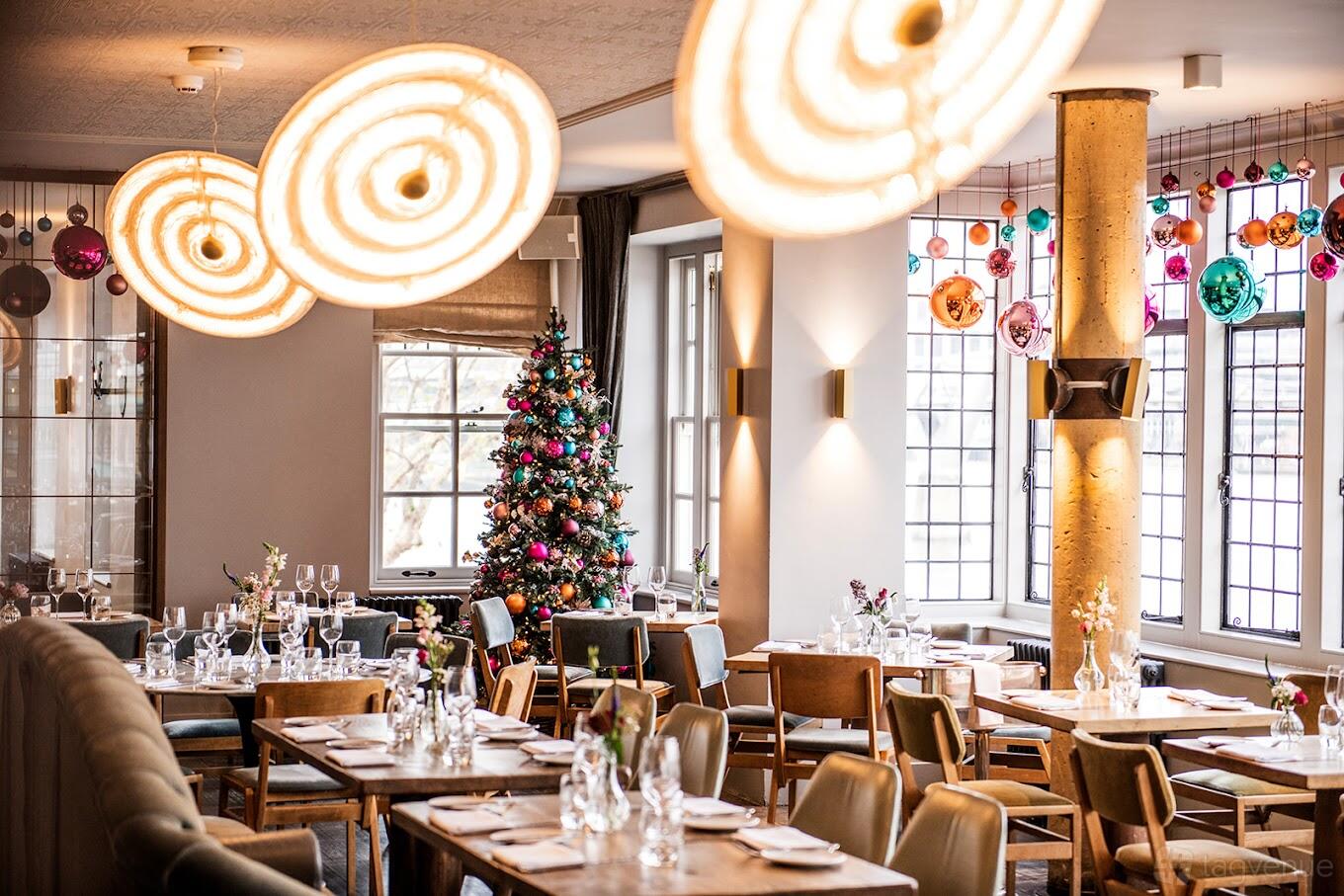 A dining room with large round pendant lights, decorated Christmas tree, and colorful glass ornaments at Swan, Shakespeare's Globe.