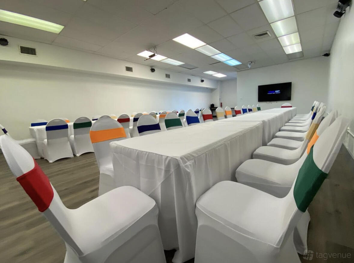 An event space with white chairs featuring colorful sashes, long tables with white linens, and bright ceiling lights at Washington's Events.
