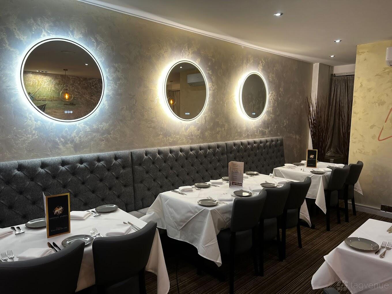 An event space in a restaurant with tufted banquette seating, round backlit mirrors, and tables set with white linens at Little India - Walsall.