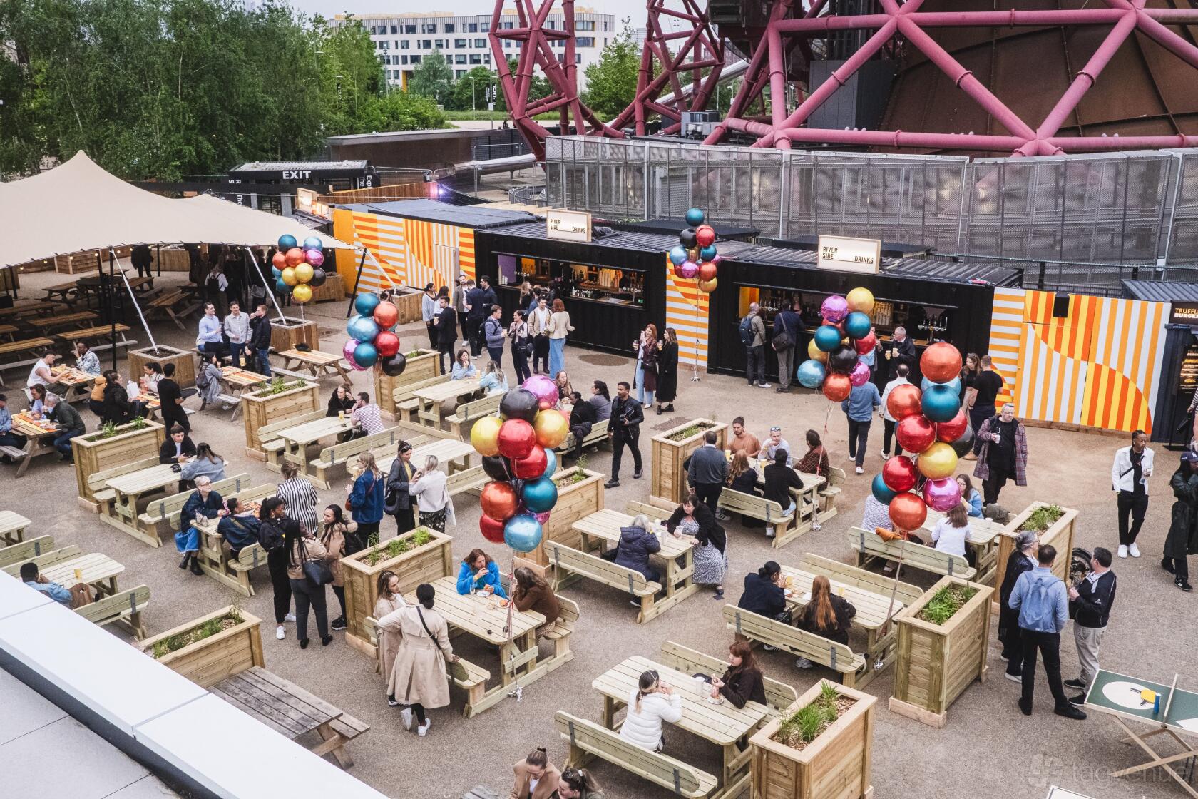 An outdoor restaurant space with wooden picnic tables, striped vendor stalls, and colorful balloon decorations at Riverside East.