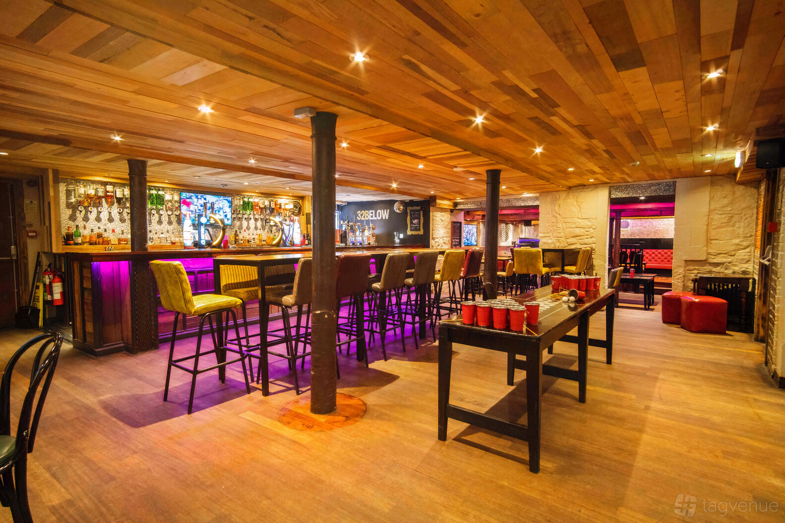 A basement bar with a wooden ceiling, purple-lit bar counter, and high stools at 32Below.