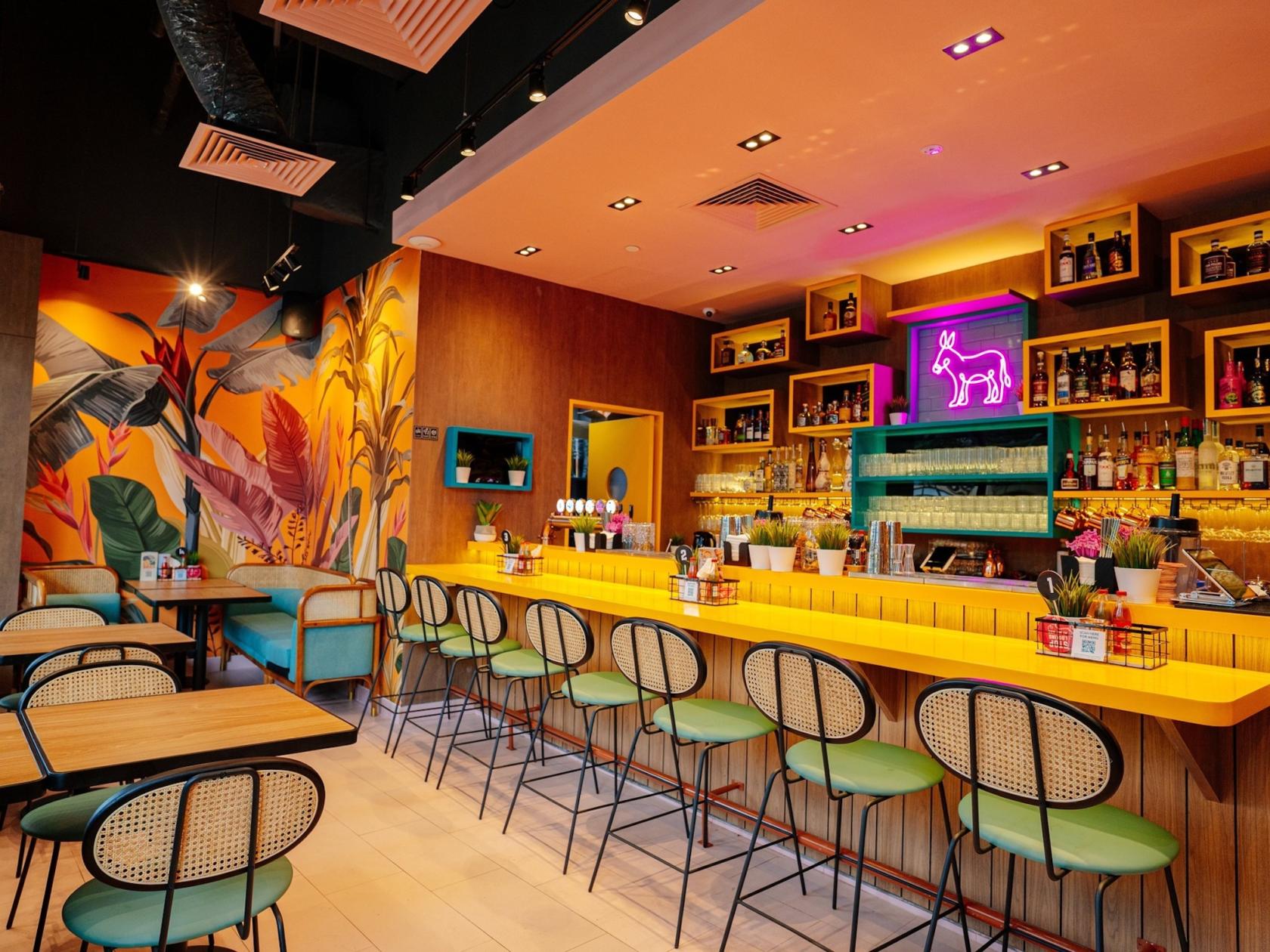 A restaurant with a vibrant mural, yellow bar counter, neon sign, and green bar stools at Chimichanga Holland Village.
