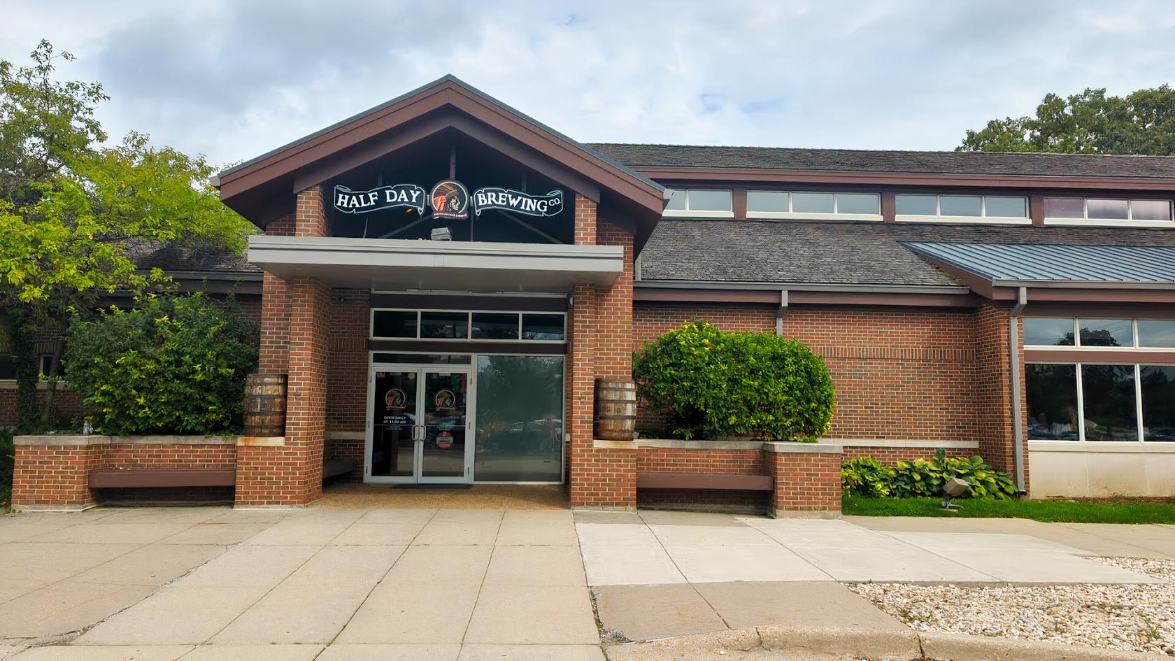 A brick pub with a covered entrance, large glass doors, and decorative barrels at Half Day Brewing.