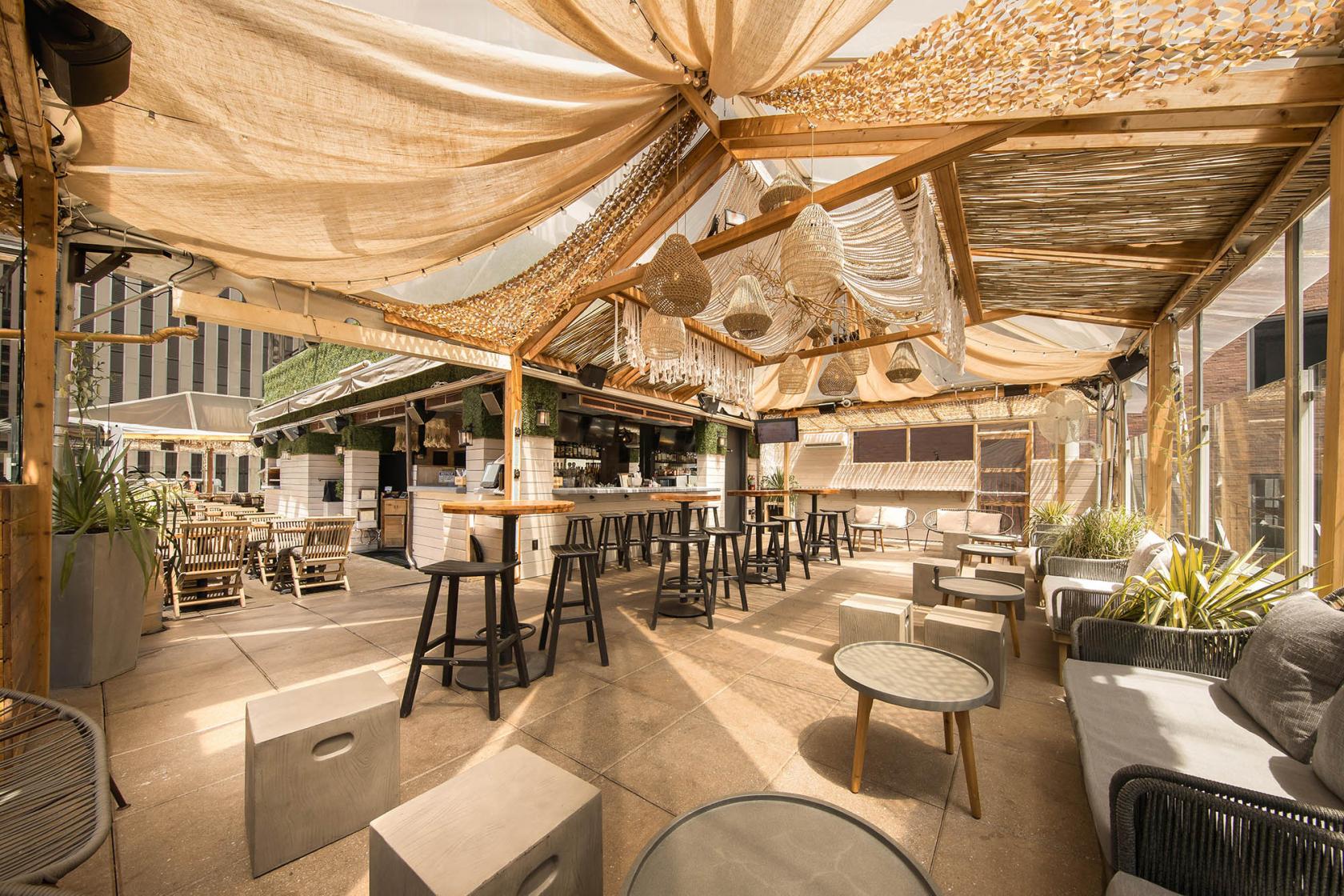A rooftop bar with wood beams, draped fabric ceiling, hanging basket lights, and high-top seating at Haven Rooftop.