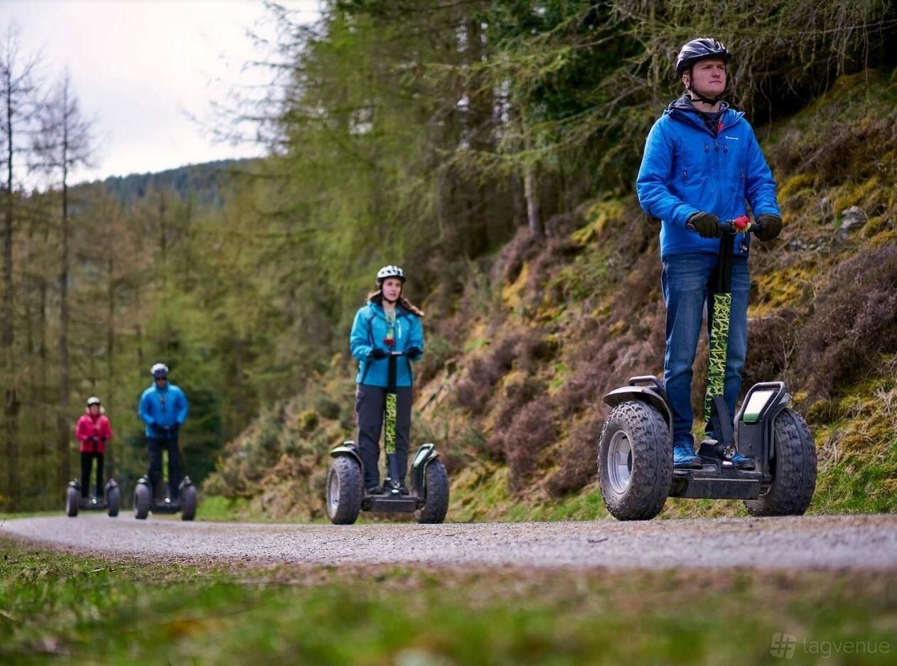 An outdoor event venue with people riding Segways on a gravel path surrounded by tall pine trees at Go Ape Whinlatter.