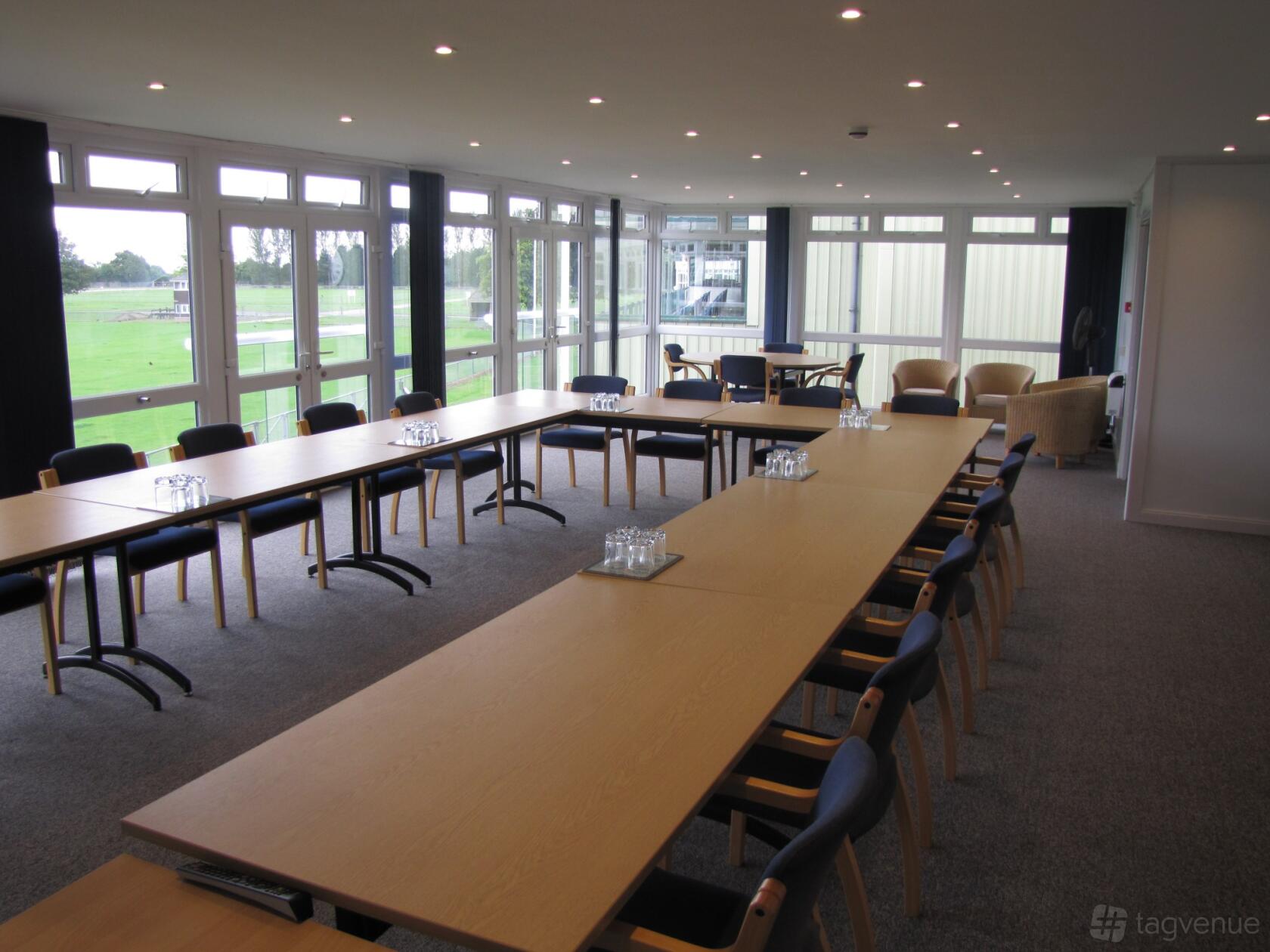 A meeting room with large windows, U-shaped wooden tables, black chairs, and natural lighting at South Of England Event Centre.