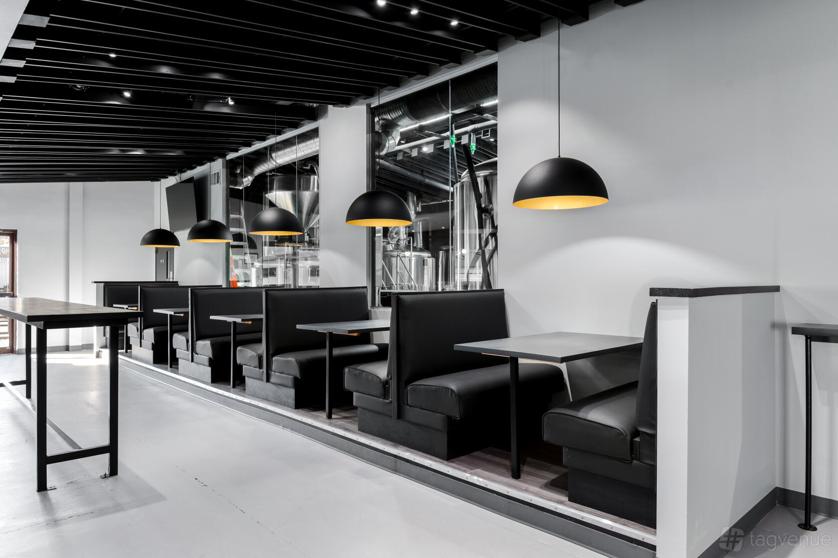 An event space with black booth seating, pendant lighting, and an open view to brewing tanks at Mutation Brewing Company.