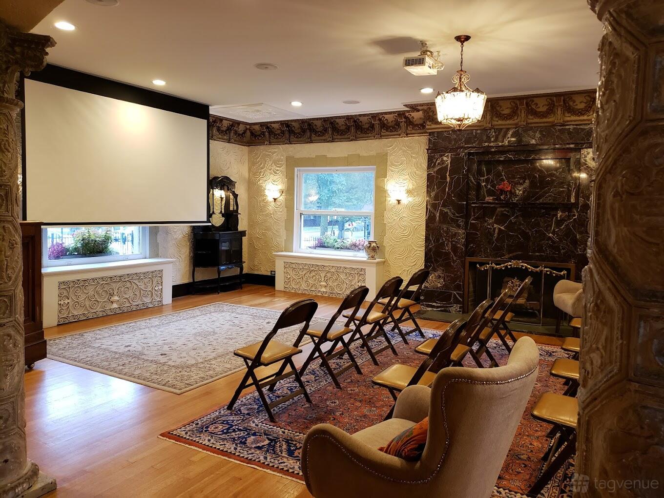 An event venue parlor with ornate molding, a marble fireplace, chandelier lighting, and folding chairs at Colvin House.