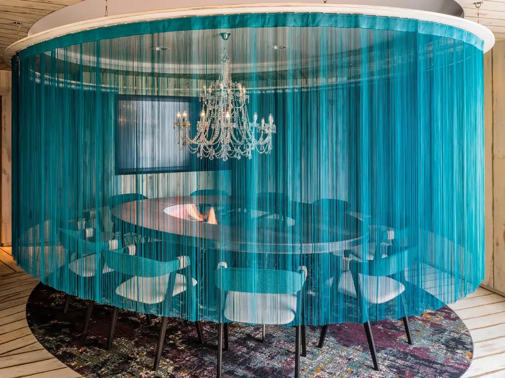 A hotel meeting room with a round table, white chairs, blue sheer curtain divider, and crystal chandelier at Novotel Canary Wharf.
