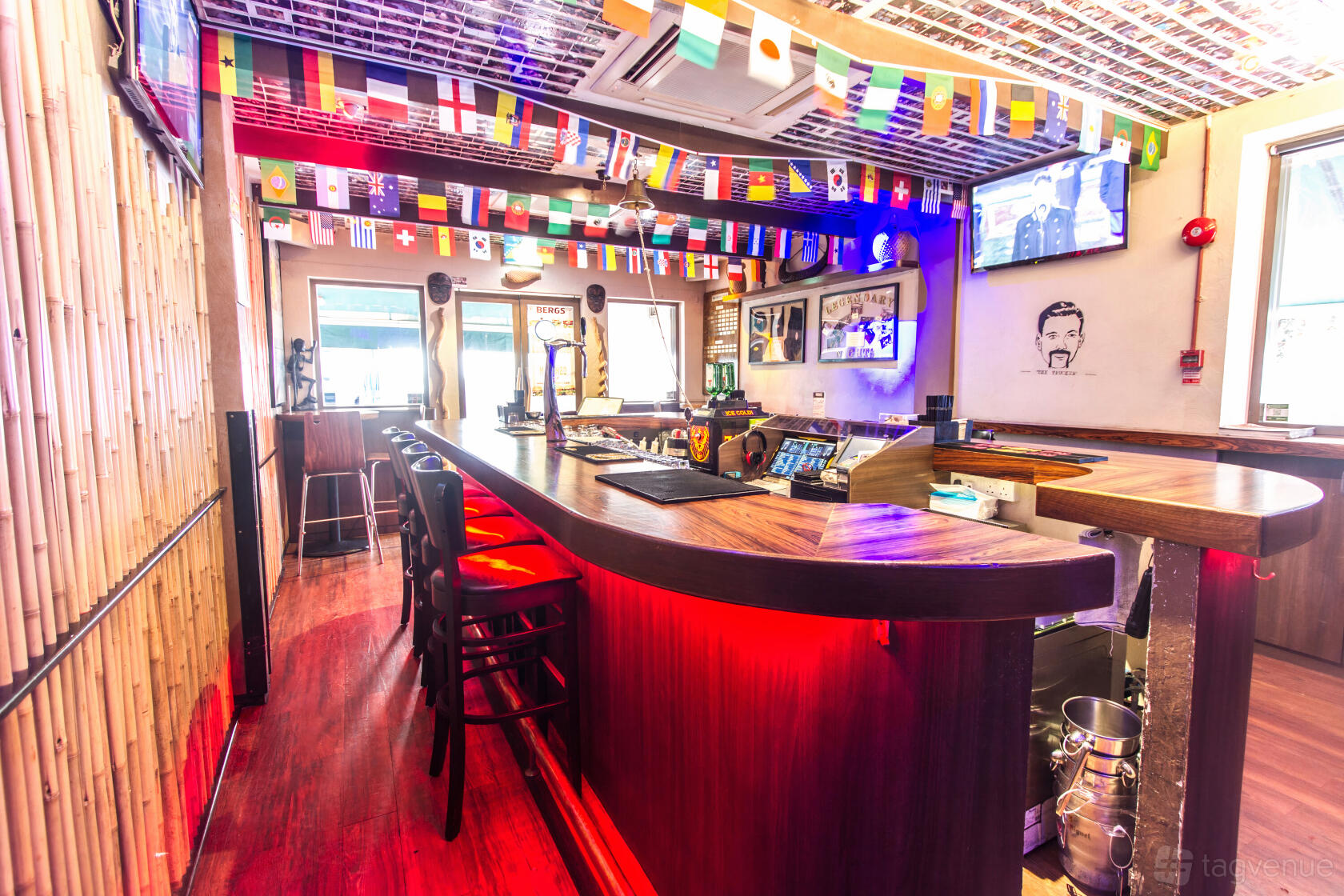 An island-style bar with a wood counter, bamboo walls, international flags, and bar stools at Mogambo Bar & Restaurant.