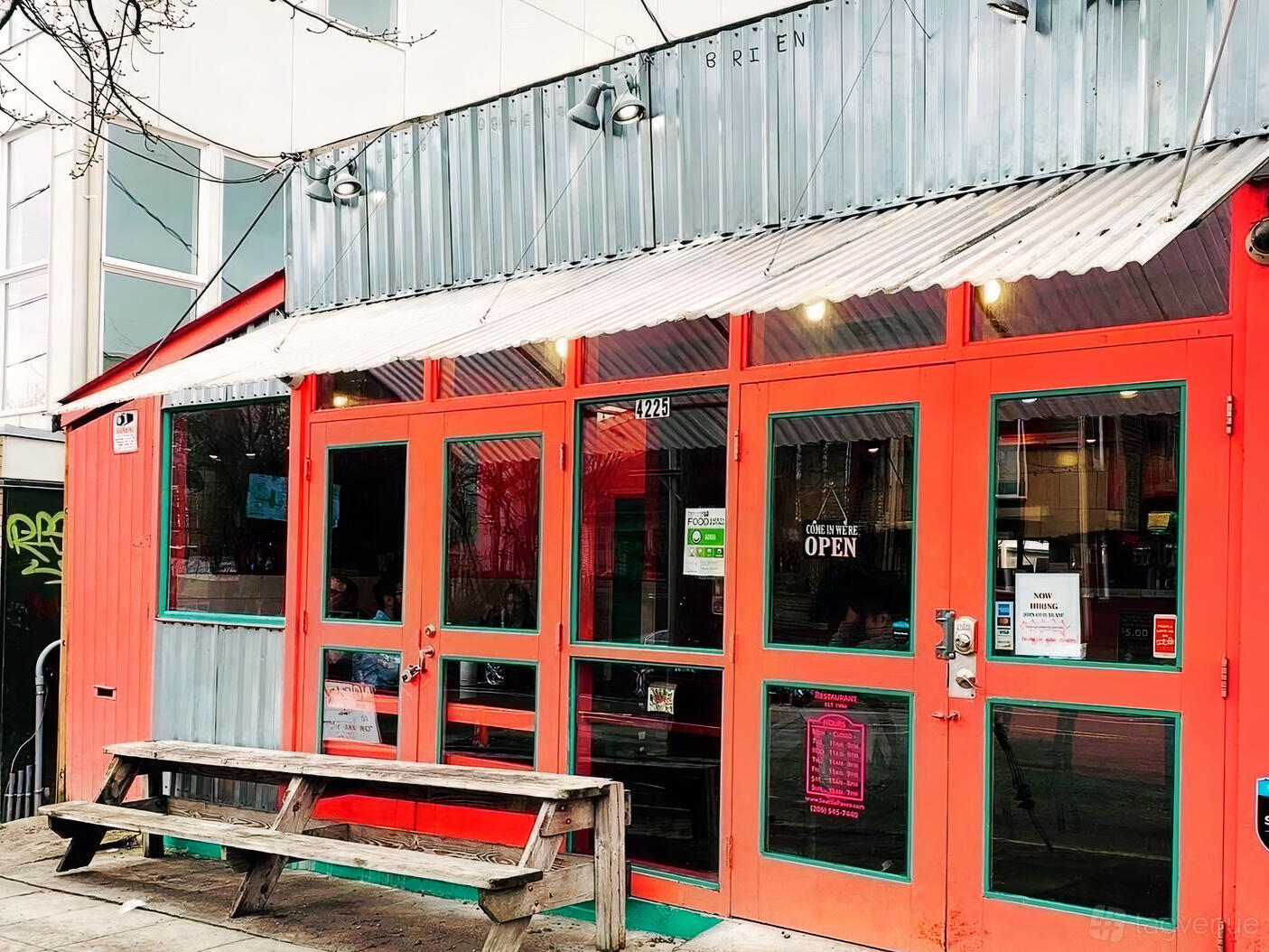 A restaurant exterior with red-framed glass doors, corrugated metal siding, and a wooden bench at Paseo.