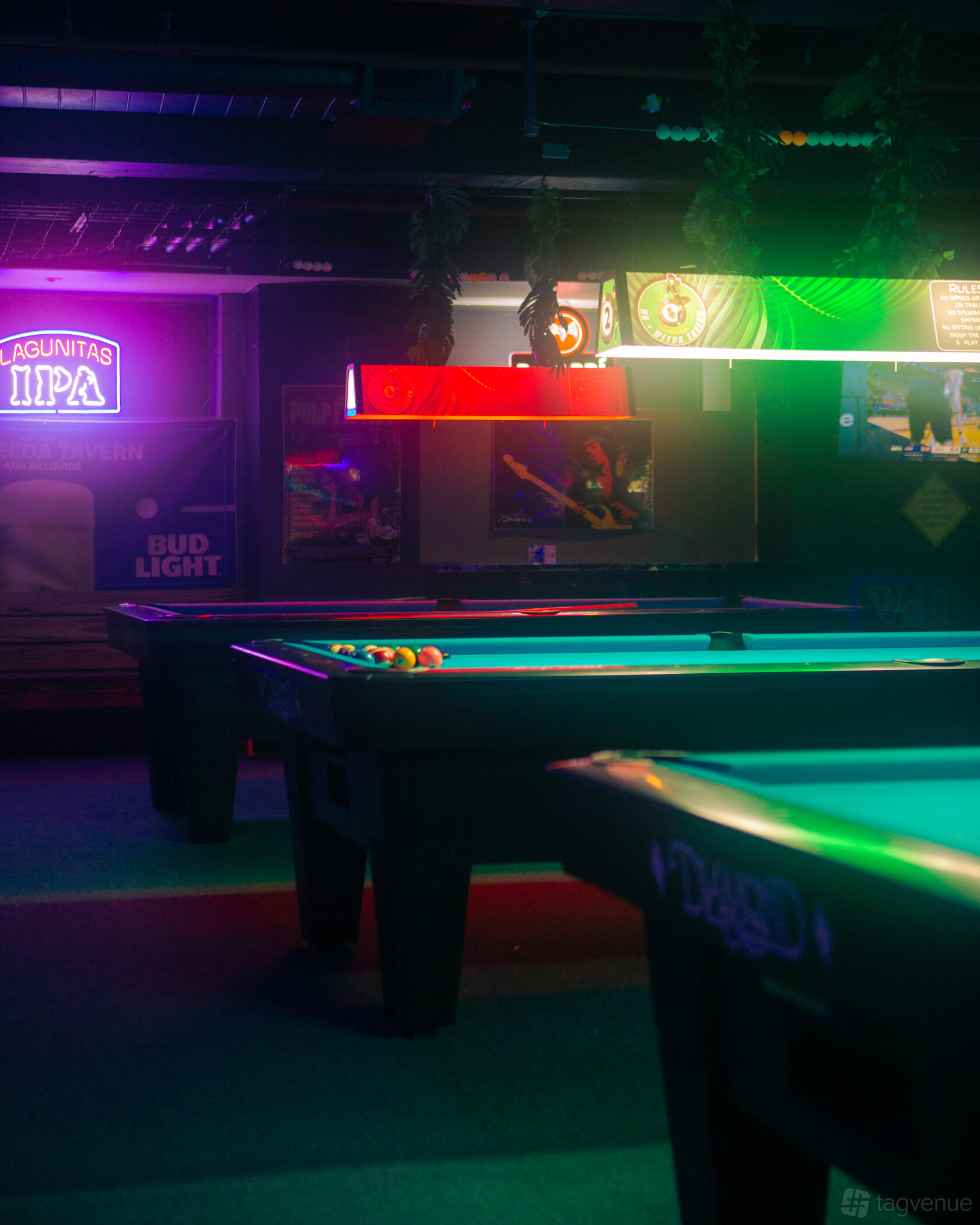 An event space in a bar with multiple pool tables, colorful overhead lights, and neon beer signs at Ha-Weeda Tavern.