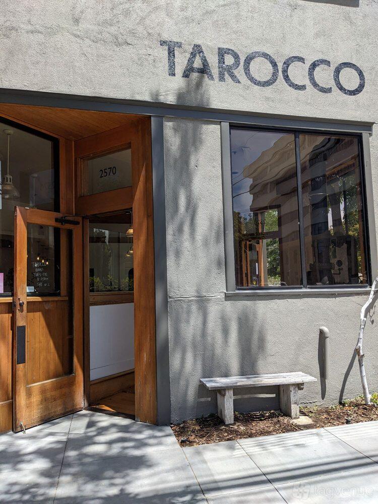A café entrance with wooden-framed glass door, large windows, and a street number sign at TAROCCO.