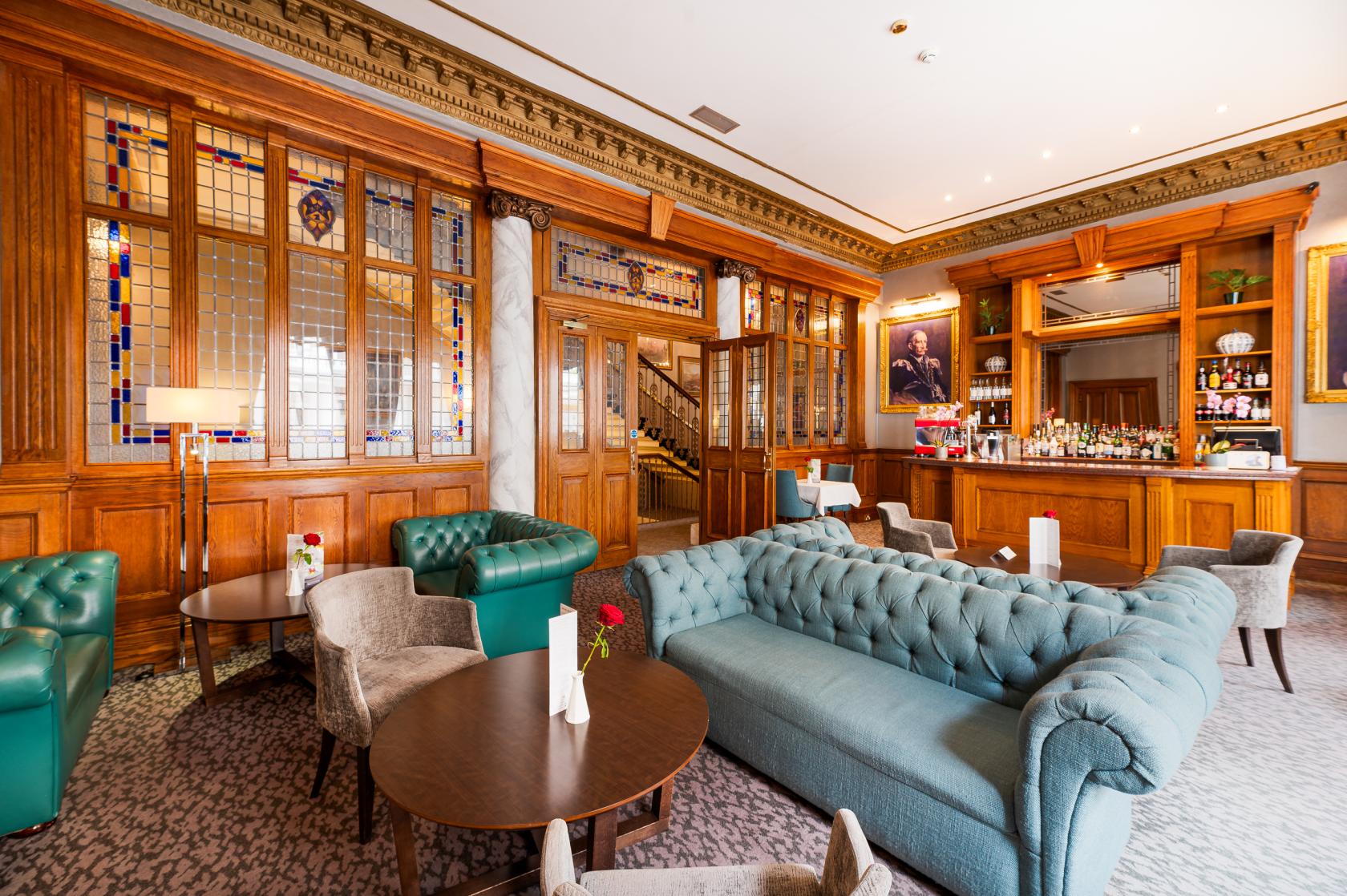 A hotel lounge with tufted teal sofas, round tables, patterned carpet, and stained glass windows at Strathmore Hotel.