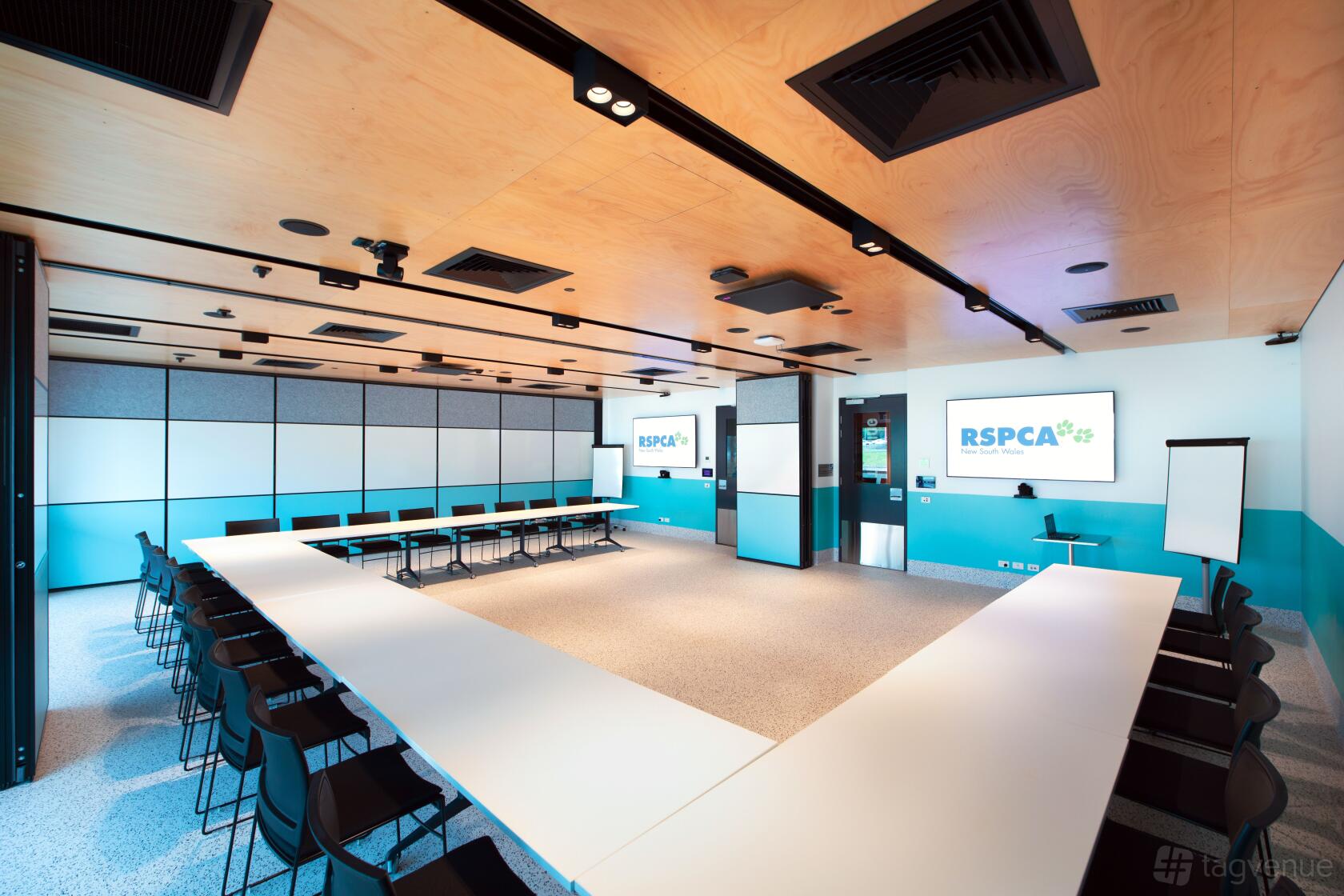 A meeting room with U-shaped white tables, black chairs, and wall-mounted screens at RSPCA NSW Sydney Education Centre.