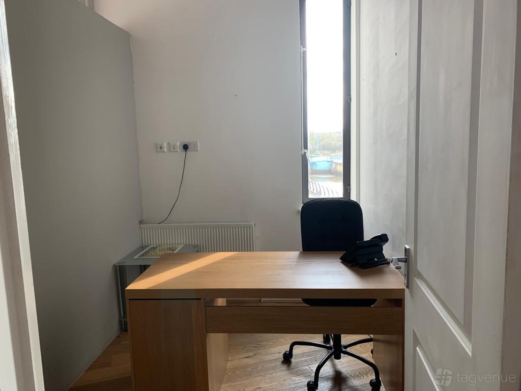 A small office with a wooden desk, black chair, and natural light from a tall window at TMH Studios.