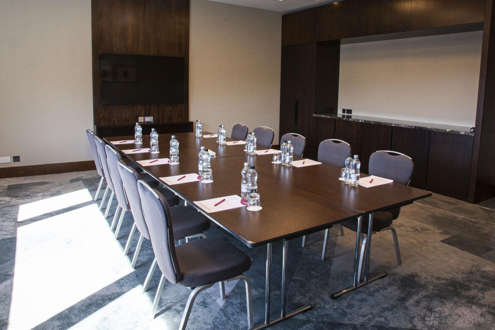 A hotel meeting room with a rectangular table, cushioned chairs, notepads, water bottles, and a wall-mounted TV at Crowne Plaza Marlow.