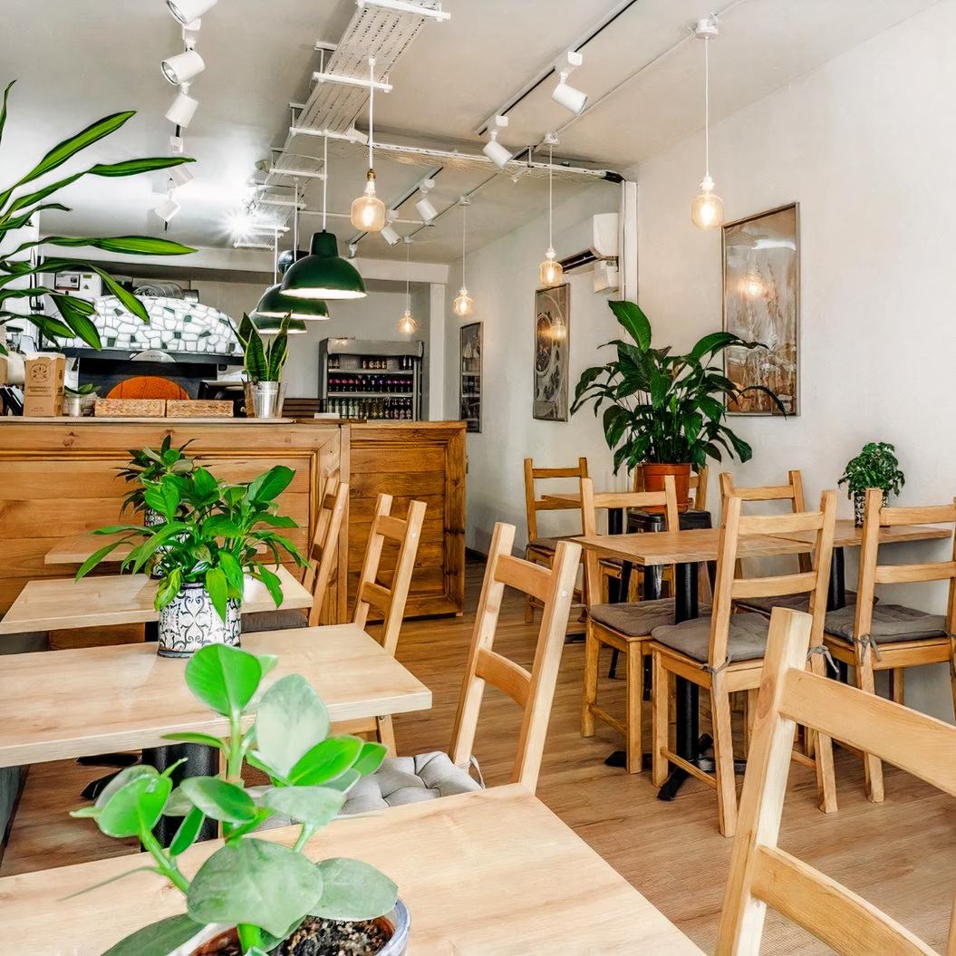 A restaurant dining area with wooden tables and chairs, potted plants, and pendant lighting at Green Mosaic Pizza & Deli.
