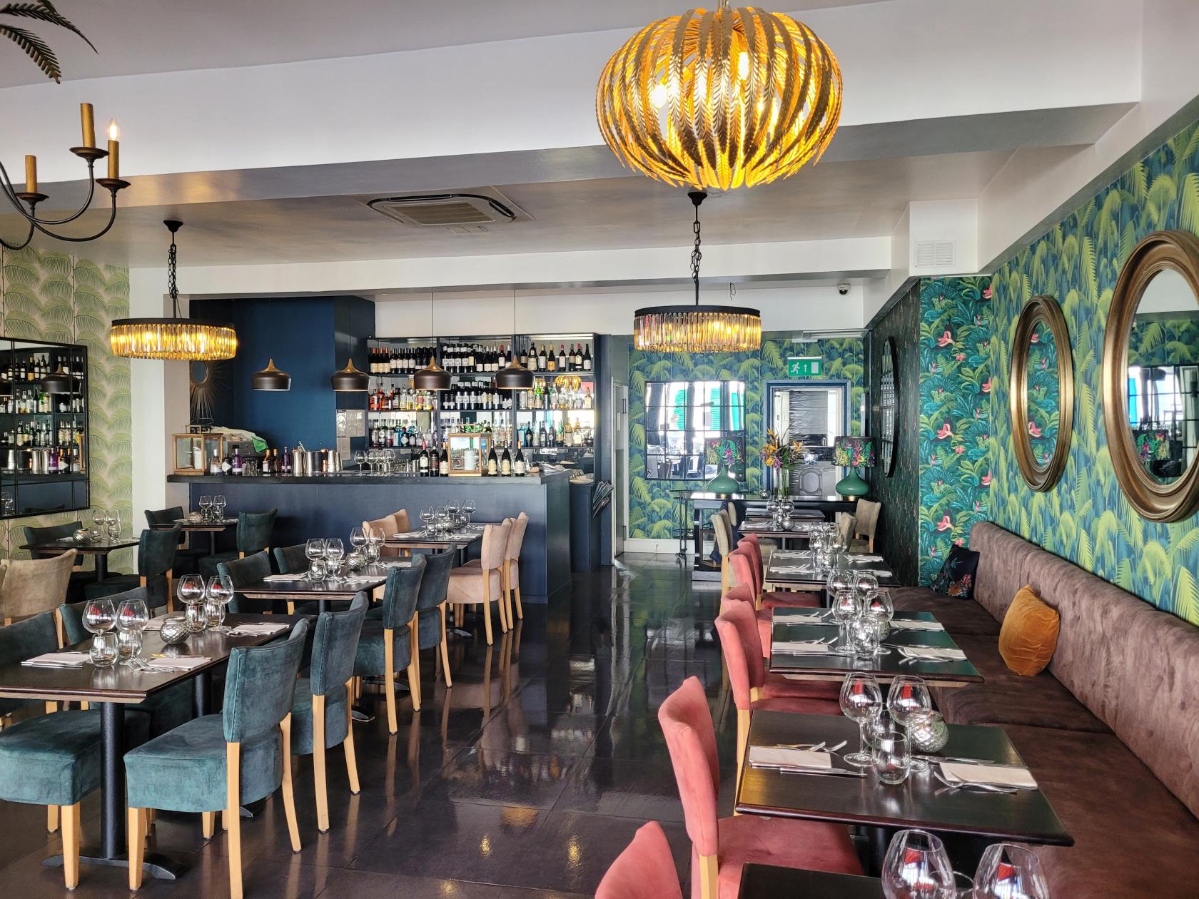 A restaurant with patterned green wallpaper, velvet chairs, round mirrors, and pendant lighting at 44 Sheen Lane.