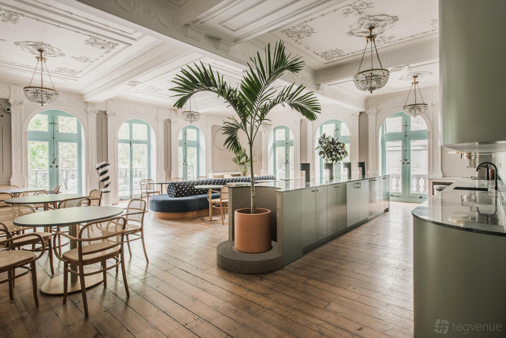 A coworking terrace with arched windows, chandeliers, potted plants, and light wood seating at Huckletree Public Hall.