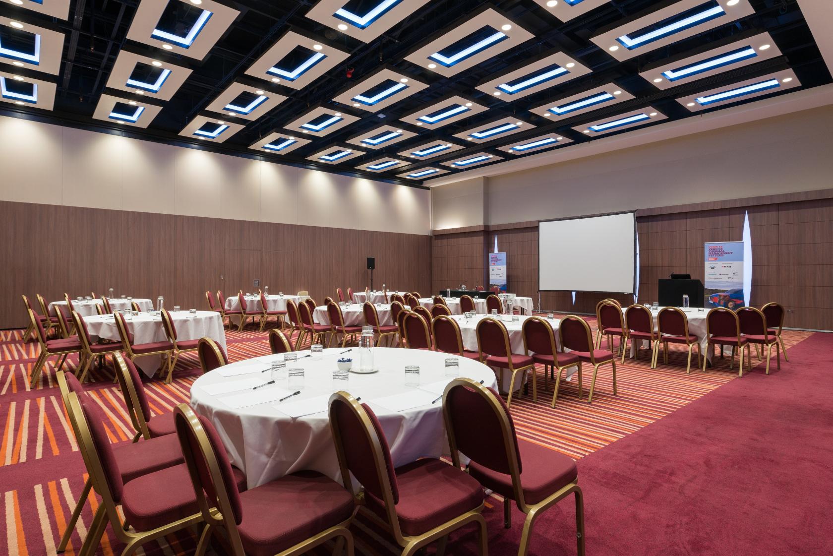 An event space with round tables, red cushioned chairs, and a projector screen at Mercure London Earl's Court.