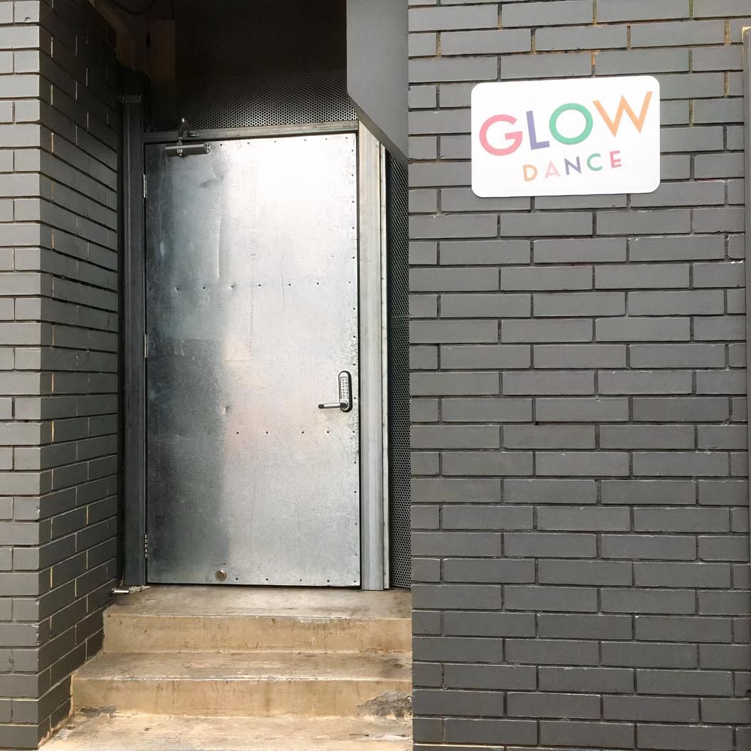 A dance studio entrance with a metal door and a Glow Dance sign above a dark brick wall at Glow Dance.