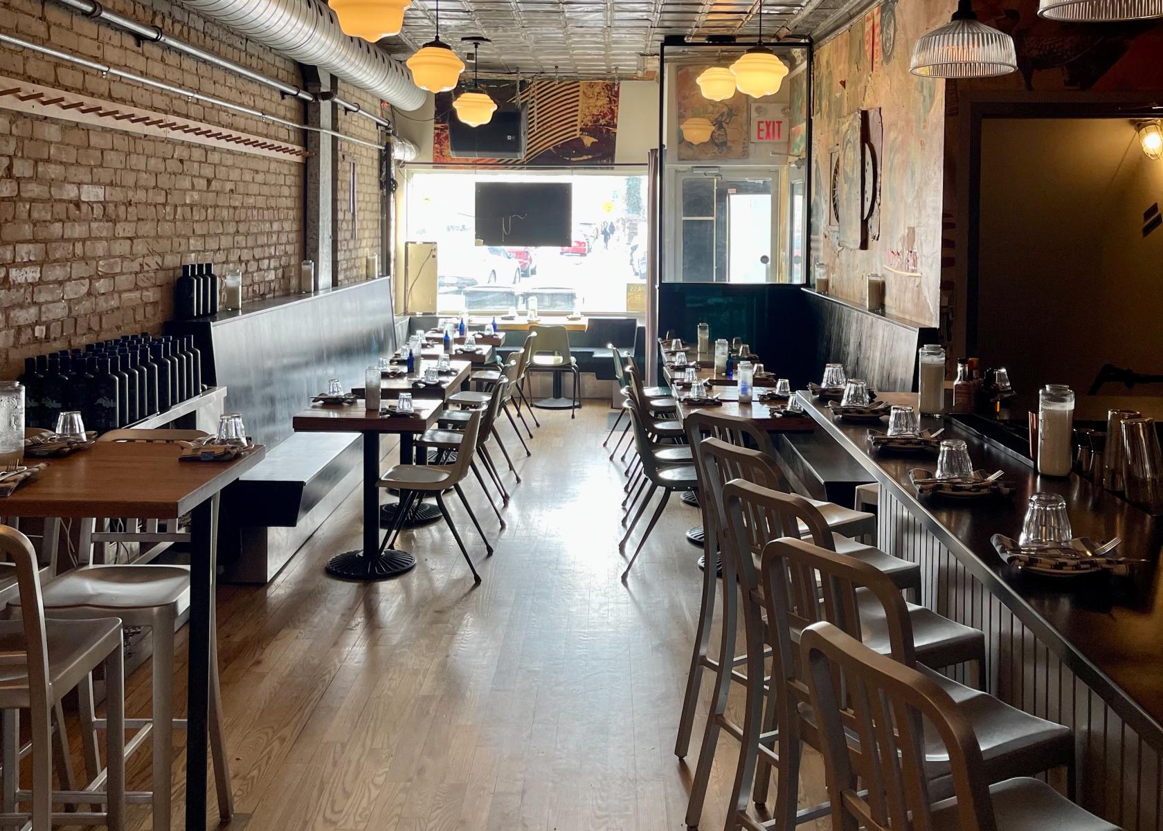 A dining room in a bar with exposed brick walls, wooden floors, and long communal tables at Bar Neon.
